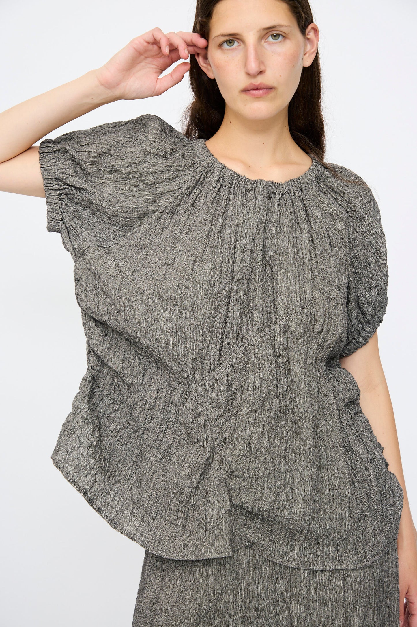 A person wearing the Lauren Manoogian Shirred Top in Charcoal, featuring a relaxed fit and gathered details, stands against a plain white background.