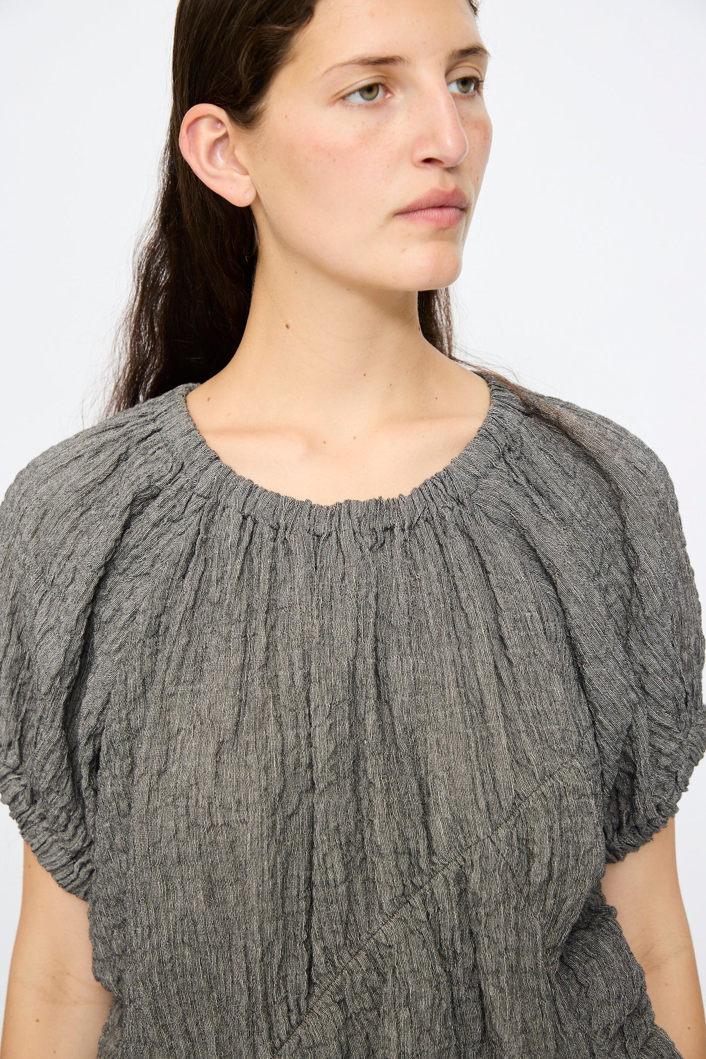 A person with long brown hair models the Lauren Manoogian Shirred Top in Charcoal, a relaxed fit short-sleeve piece, while looking slightly to the side against a plain background.