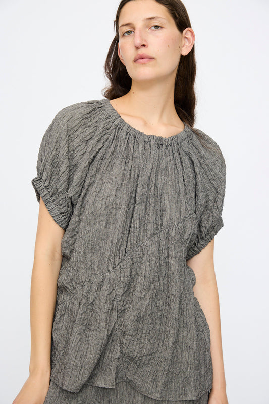 A woman with long brown hair wears the Lauren Manoogian Shirred Top in Charcoal, a relaxed fit, textured linen-blend short-sleeve style, as she stands against a plain white background.
