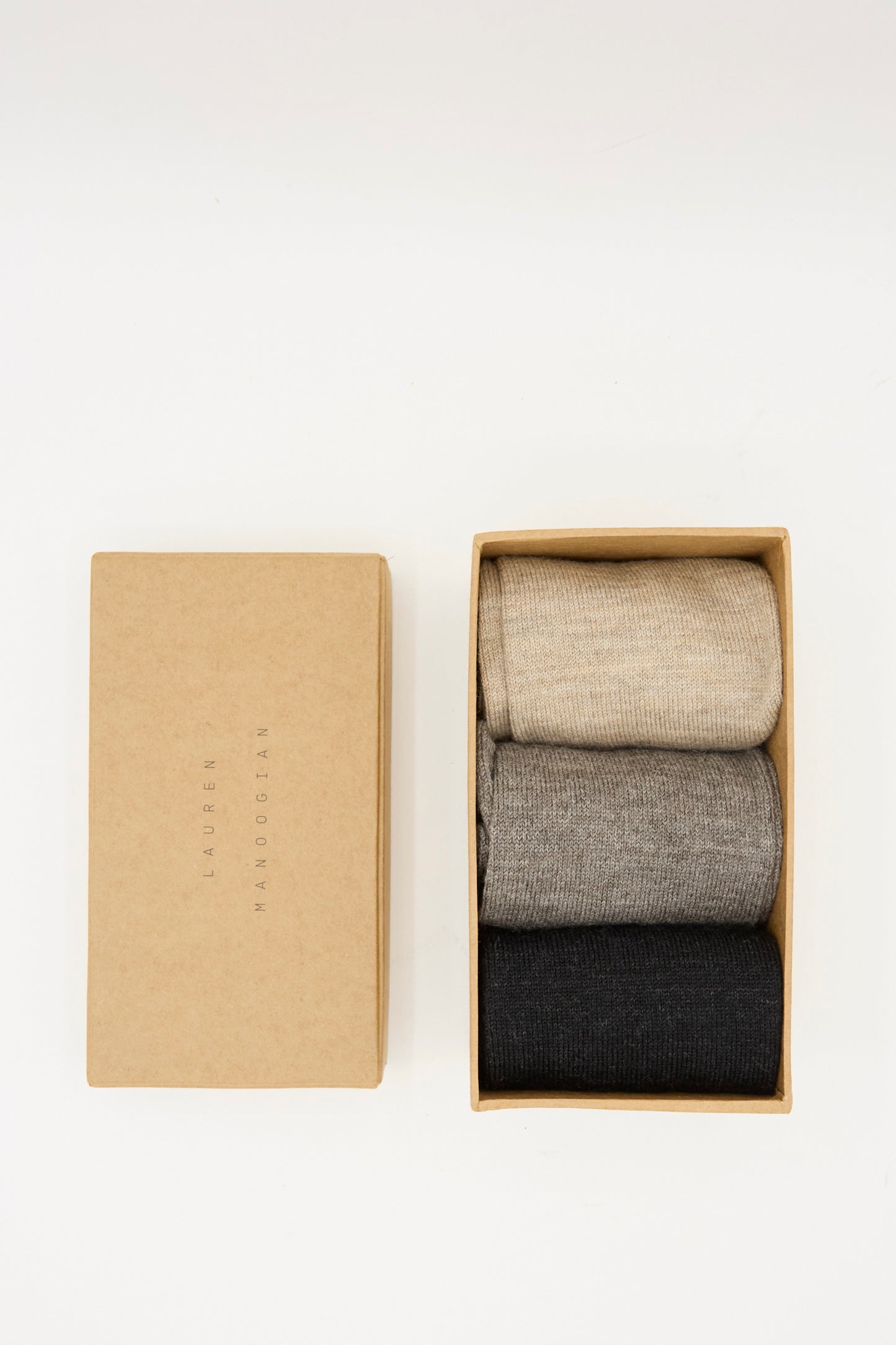 A Lauren Manoogian Sock Set in Mix: three pairs of mid-calf socks in beige, gray, and black, displayed in a cardboard box with its lid beside it on a white background.