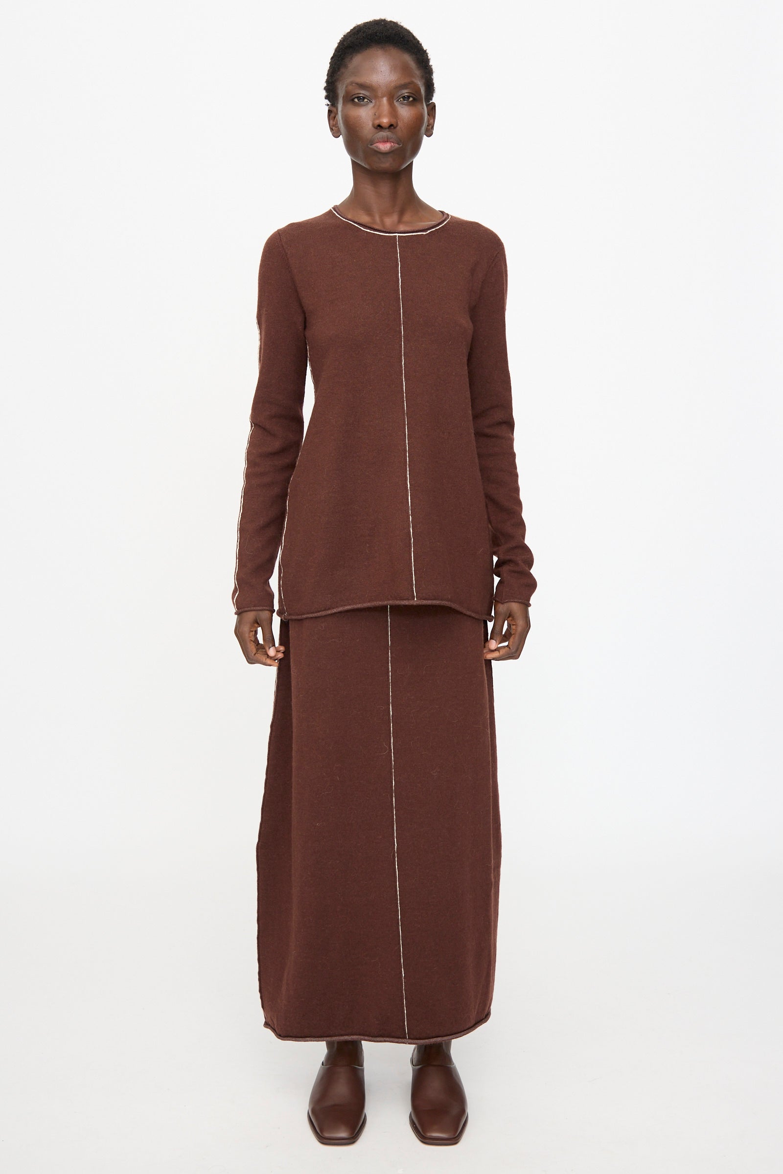 A person faces forward against a plain white background, wearing the Lauren Manoogian Stitch Crewneck in Merlot, paired with a matching long skirt featuring visible front seams, and brown shoes.