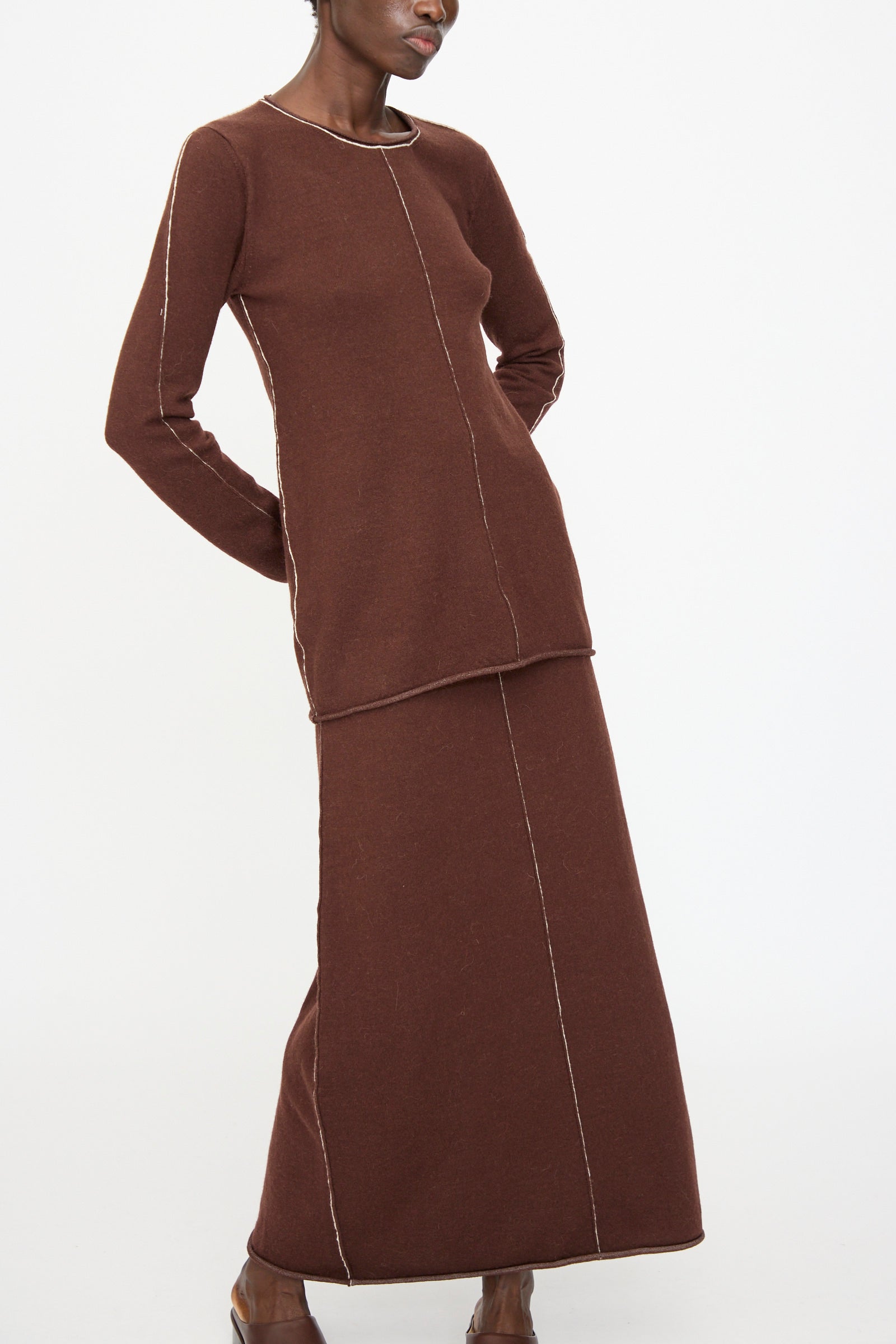 A person wears the Lauren Manoogian Stitch Crewneck in Merlot, a long brown knit dress with white seams and a fitted top, styled over a slim-fit alpaca blend shirt, standing against a plain white background.