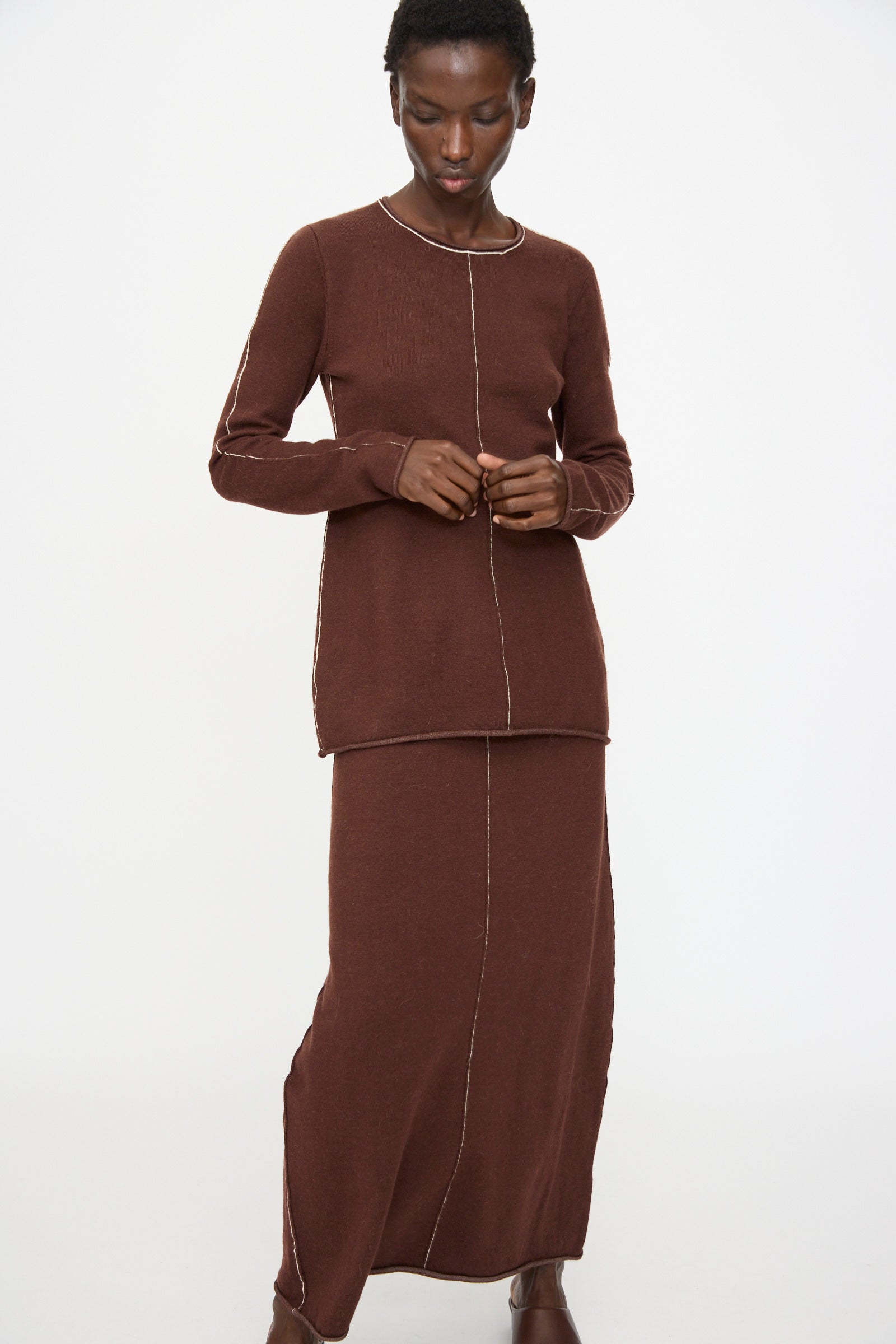 A person wears the Lauren Manoogian Stitch Crewneck in Merlot, a slim-fit long sleeve knit top with visible white seams, styled with a matching long skirt, against a plain white background.