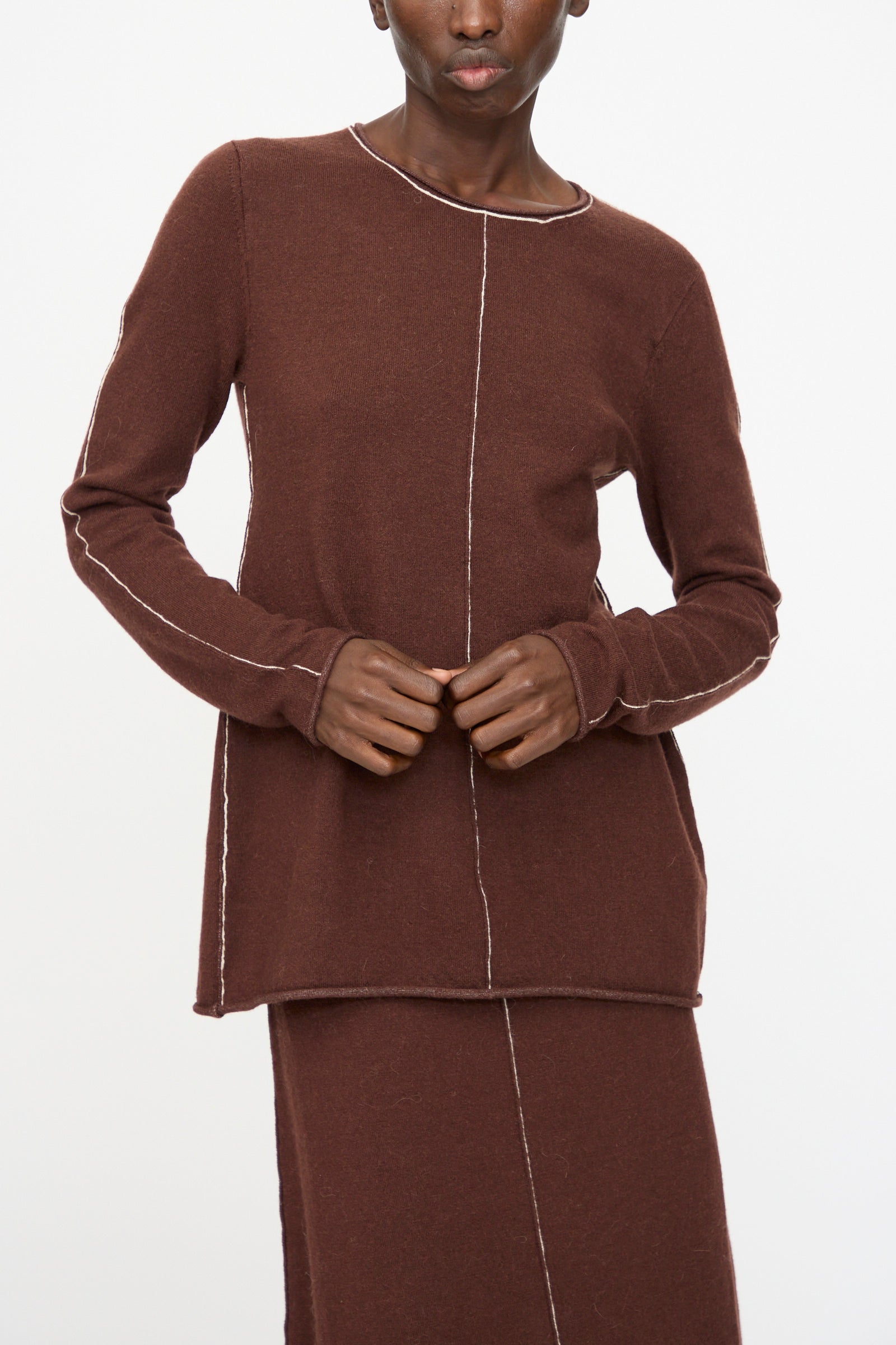 A person wearing the Lauren Manoogian Stitch Crewneck in Merlot, a slim fit long sleeve top with visible white seams, and a matching brown skirt stands against a plain white background.
