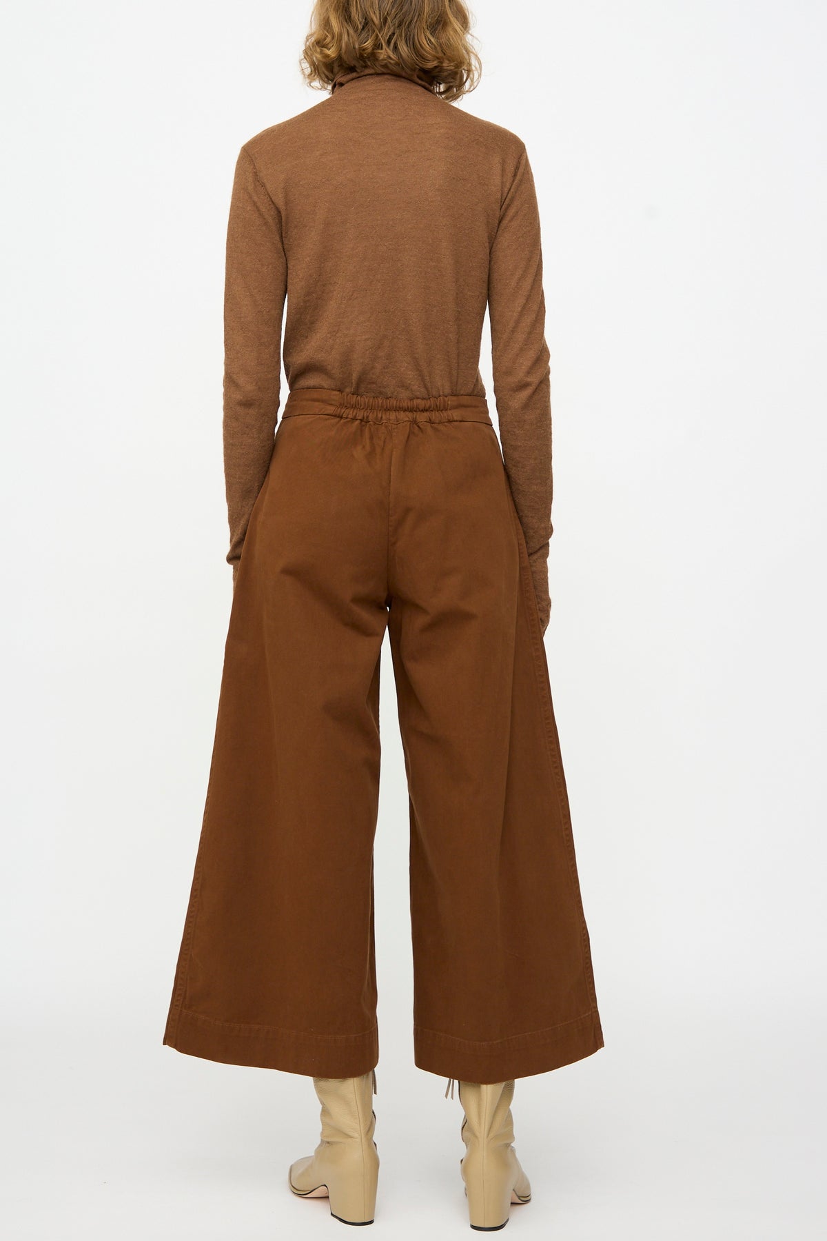 A person stands facing away, wearing a brown long-sleeve top, Lauren Manoogian Sweep Pants in Rust—a relaxed fit, wide-leg cotton style made in Peru—and beige boots, against a plain white background.