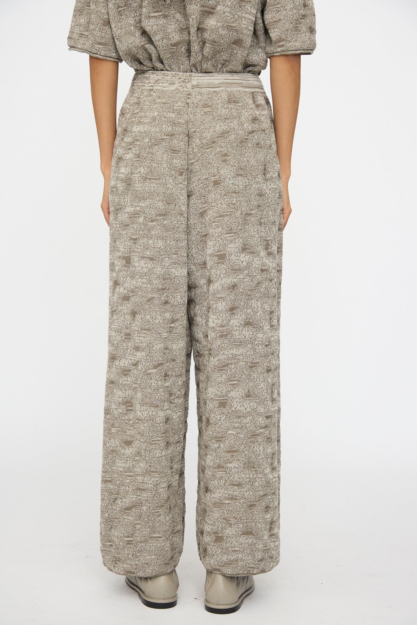 The lower half of a person is shown wearing the Lauren Manoogian Terra Pants in Rock Marl—textured, loose-fitting taupe pants with an elastic waist—paired with a matching top against a plain white background.