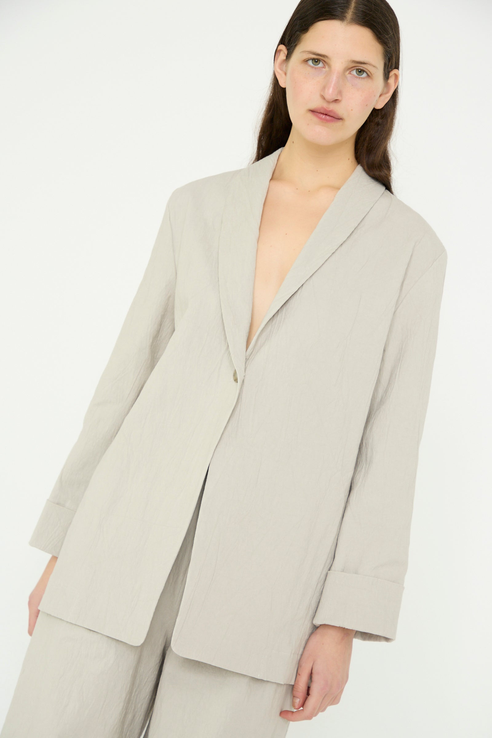 A person stands against a plain background, wearing the Texture Blazer in Pewter by Lauren Manoogian with a shawl collar and matching pants, looking forward.