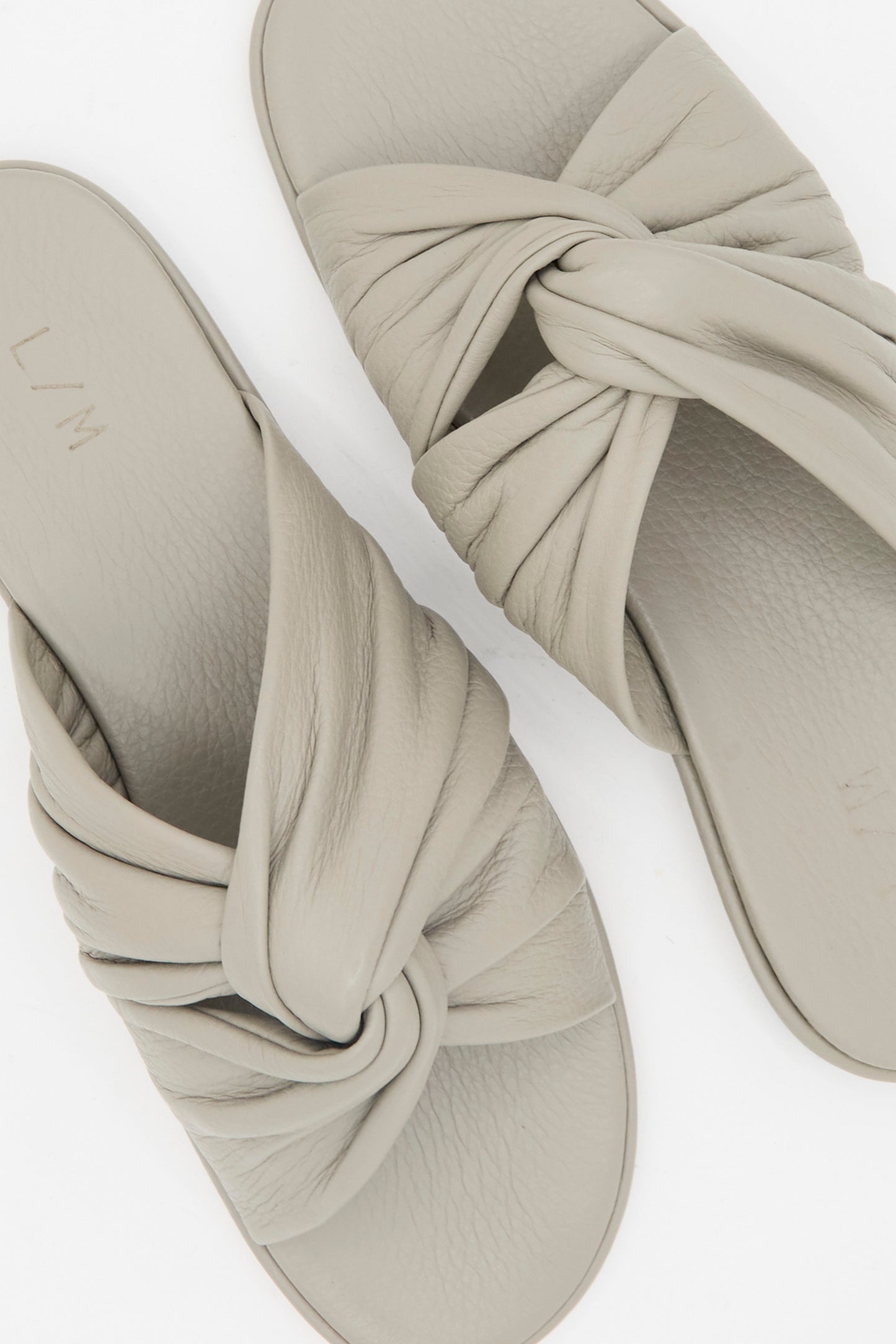 The Lauren Manoogian Twist Slide in Stone features light beige leather twist vamp straps and flat soles, shown against a white background.