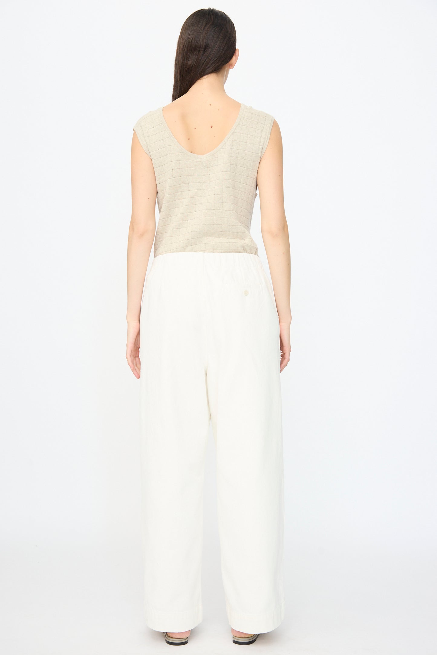 A woman faces away, wearing a sleeveless beige top and Lauren Manoogian's Washed Trouser in White—loose, relaxed fit with an elasticated waist—against a plain white background.