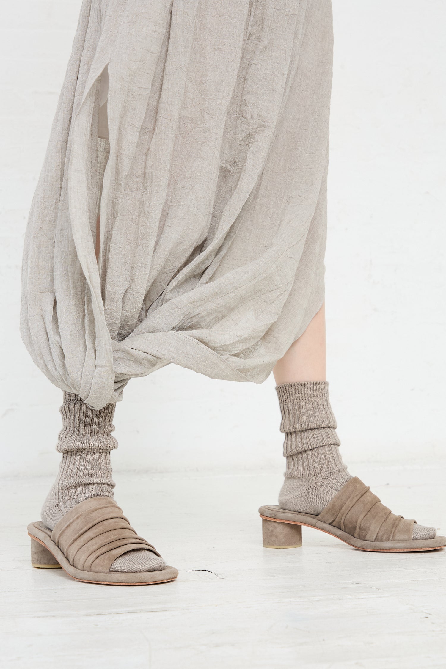 Close-up of a person's lower legs wearing beige sandals and grey socks, draped with a light grey, flowing Lauren Manoogian Gauze Twist Skirt in Ash.