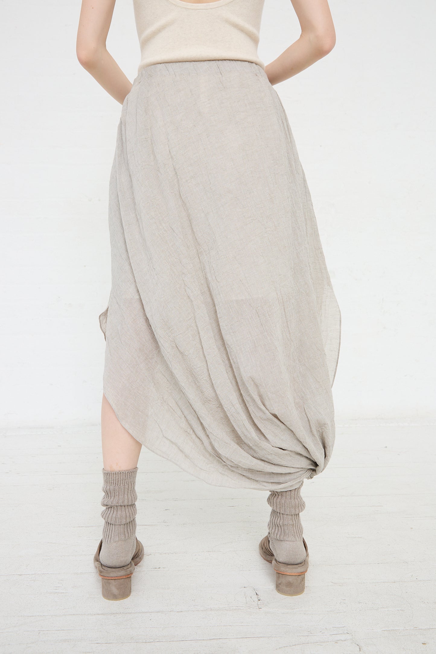 Woman viewed from behind wearing a draped beige cotton/linen blend Gauze Twist Skirt in Ash by Lauren Manoogian, grey socks, and brown clogs against a white background.