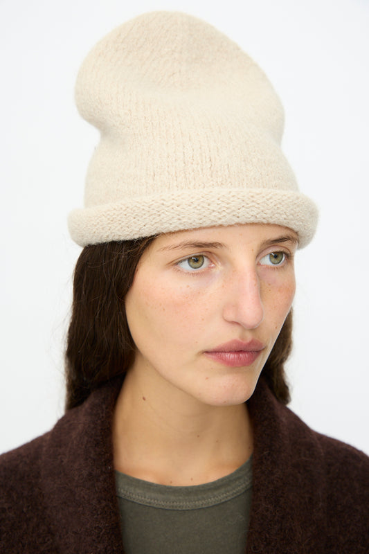 A person with long brown hair wears the Lauren Manoogian Crown Beanie in Antique Melange and a dark coat, looking slightly to the side against a plain light background.