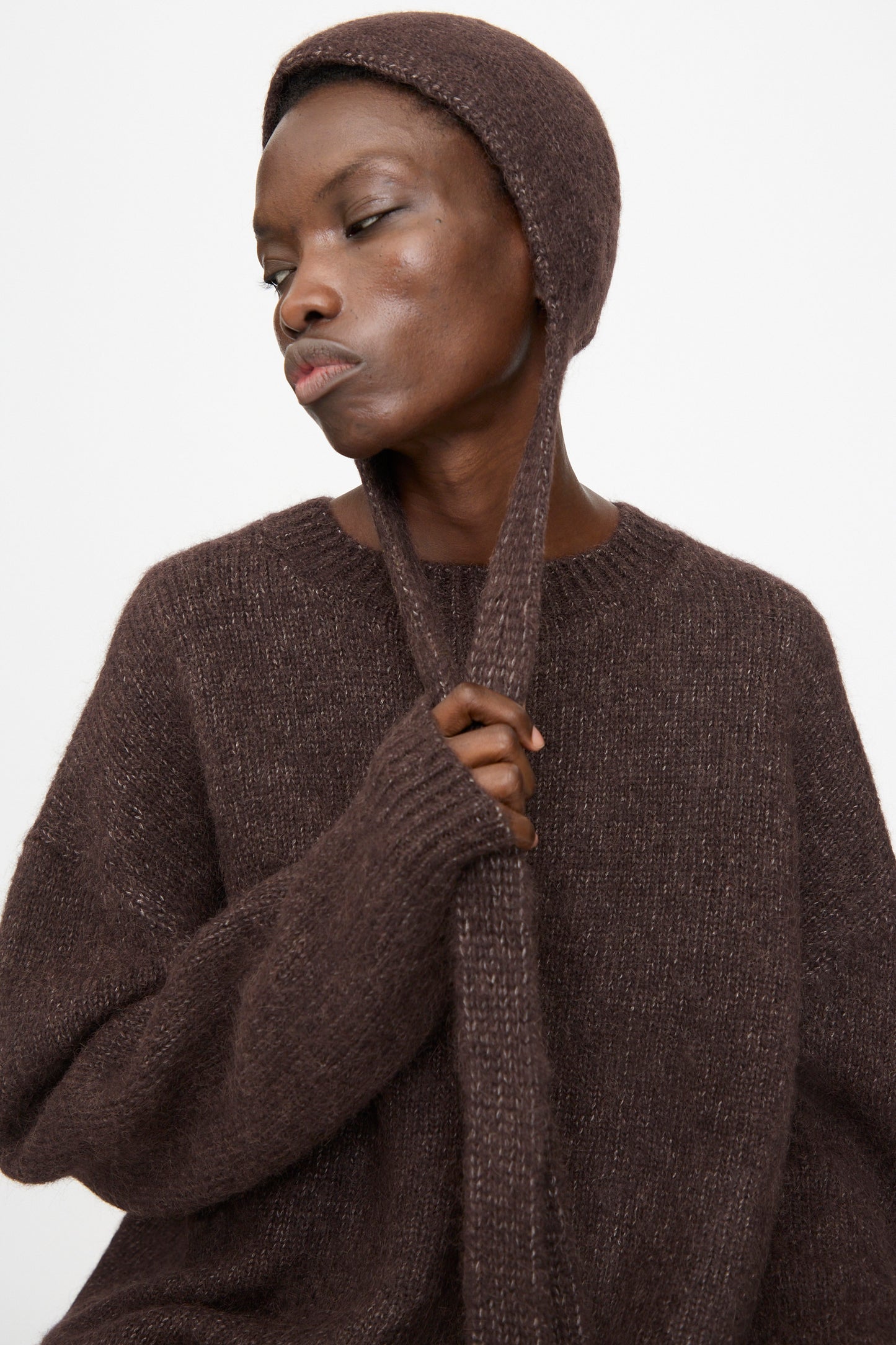 A person wears the Lauren Manoogian Loft Bonnet in Soil, holding its ties, against a plain white background.