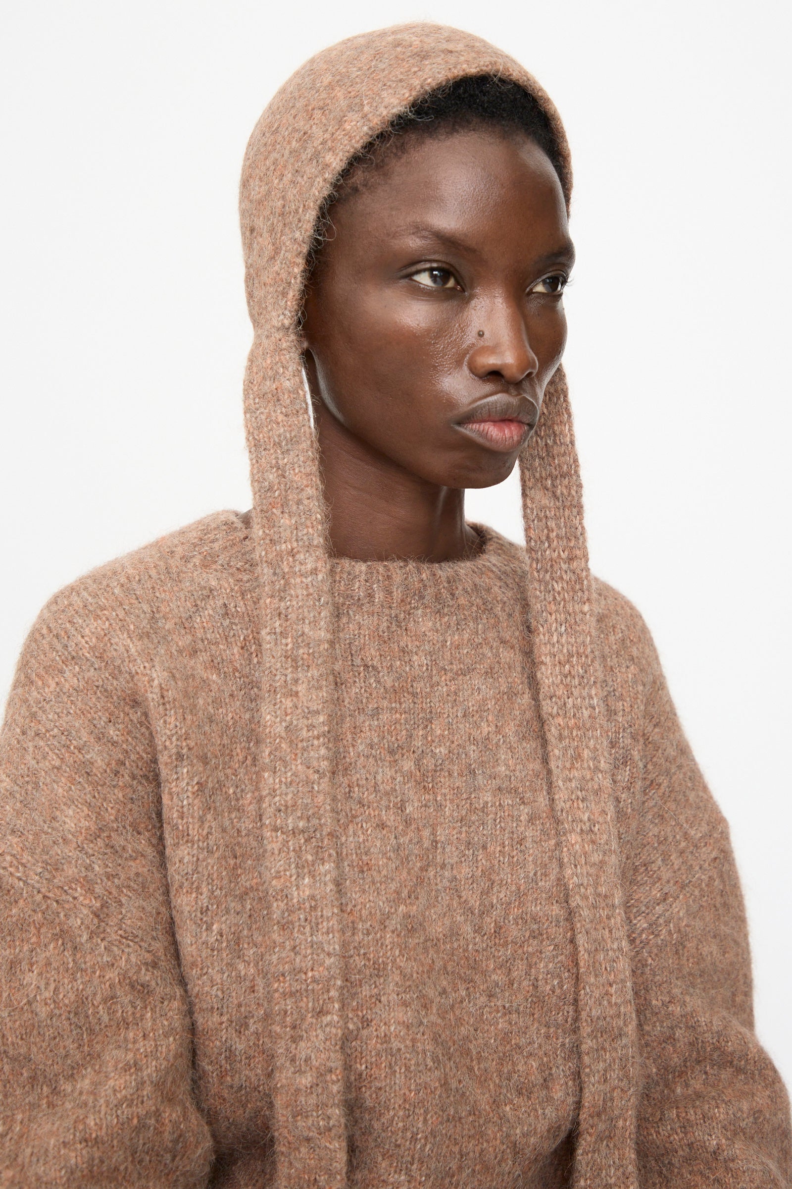 Model wears the Lauren Manoogian Loft Bonnet in Spice, a fitted bonnet with long ties, paired with a loose-knit brown sweater, crafted from a suri alpaca blend. She looks slightly to the side against a plain white background.
