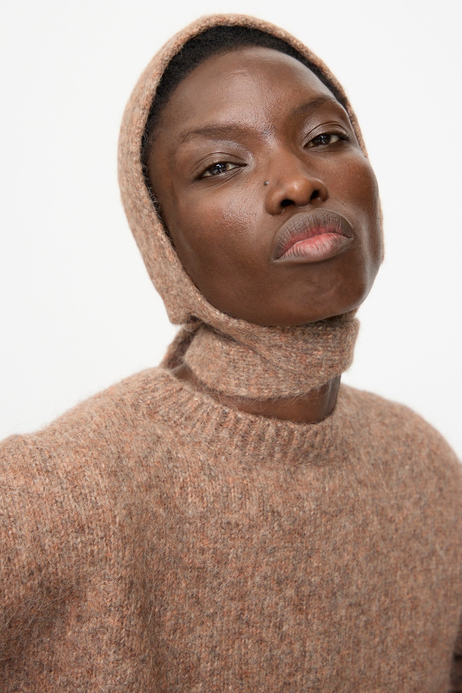 A person wears the Lauren Manoogian Loft Bonnet in Spice, paired with a light brown textured suri alpaca blend knit sweater, looking at the camera with a neutral expression against a plain background.