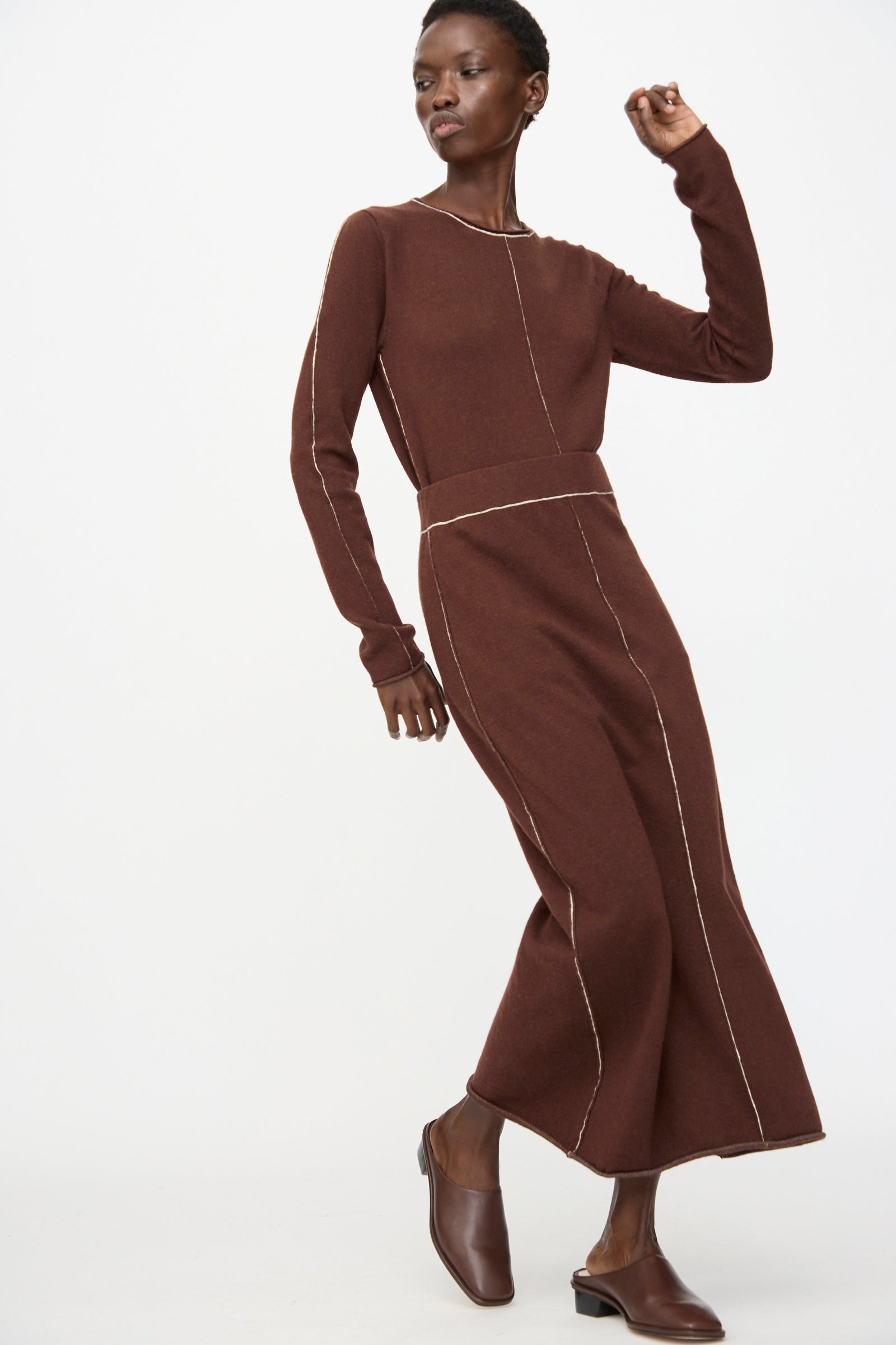 A person wears the Lauren Manoogian Stitch Skirt in Merlot, a long knit skirt with white seam details, paired with brown slip-on shoes, standing against a plain white background.