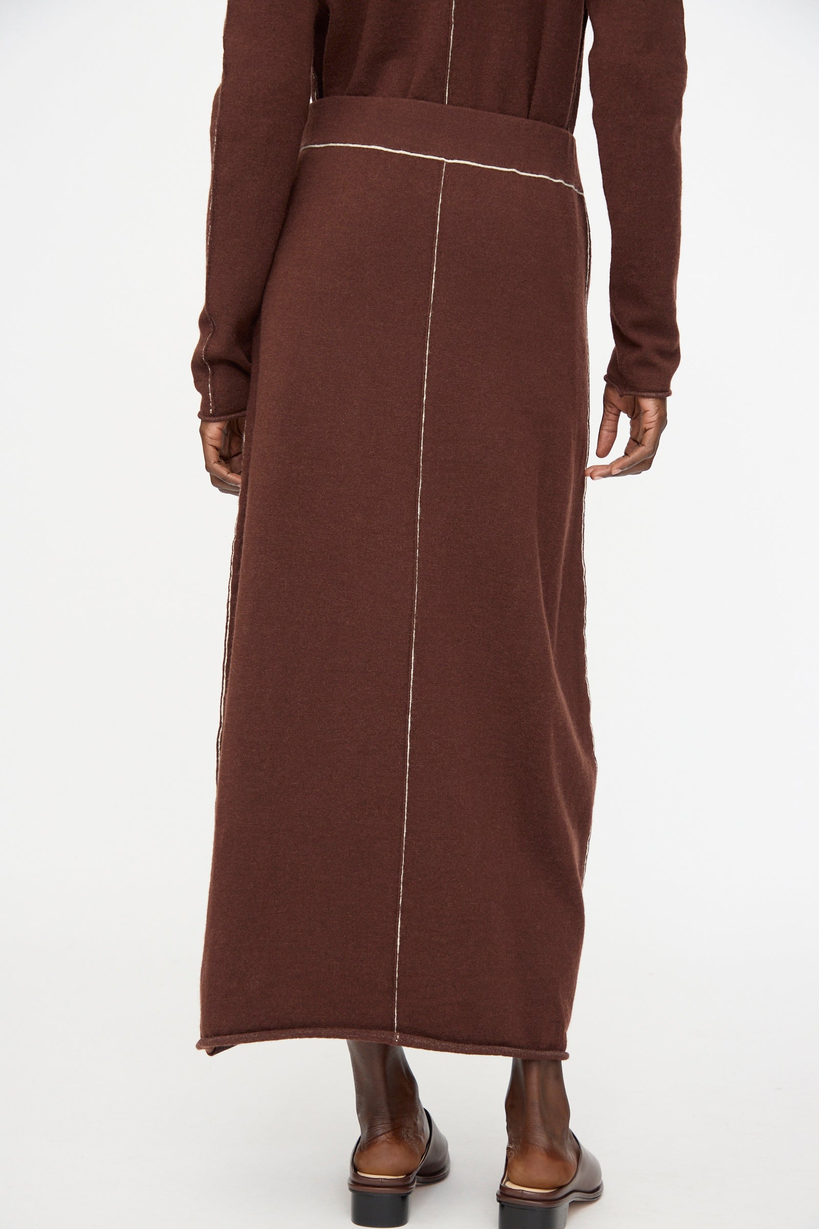 A person is shown from the back, standing on a white background, wearing Lauren Manoogian's Stitch Skirt in Merlot—a long brown maxi skirt featuring visible white stitching.
