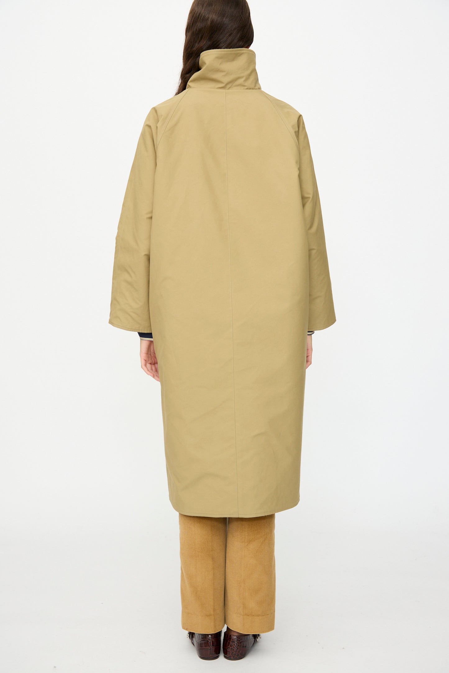 A person stands with their back to the camera, wearing the Les Prairies De Paris Dickens Moon Reversible British Trench in Taupe, paired with tan pants and brown shoes, set against a plain white background.