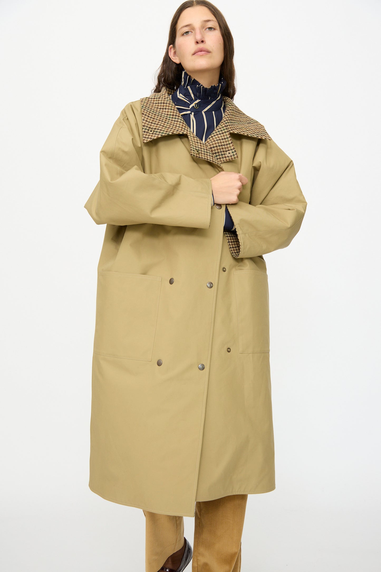 A person wears the Les Prairies De Paris Dickens Moon Reversible British Trench in Taupe, featuring a checked collar, over a navy patterned top and light brown pants, standing against a plain white background.