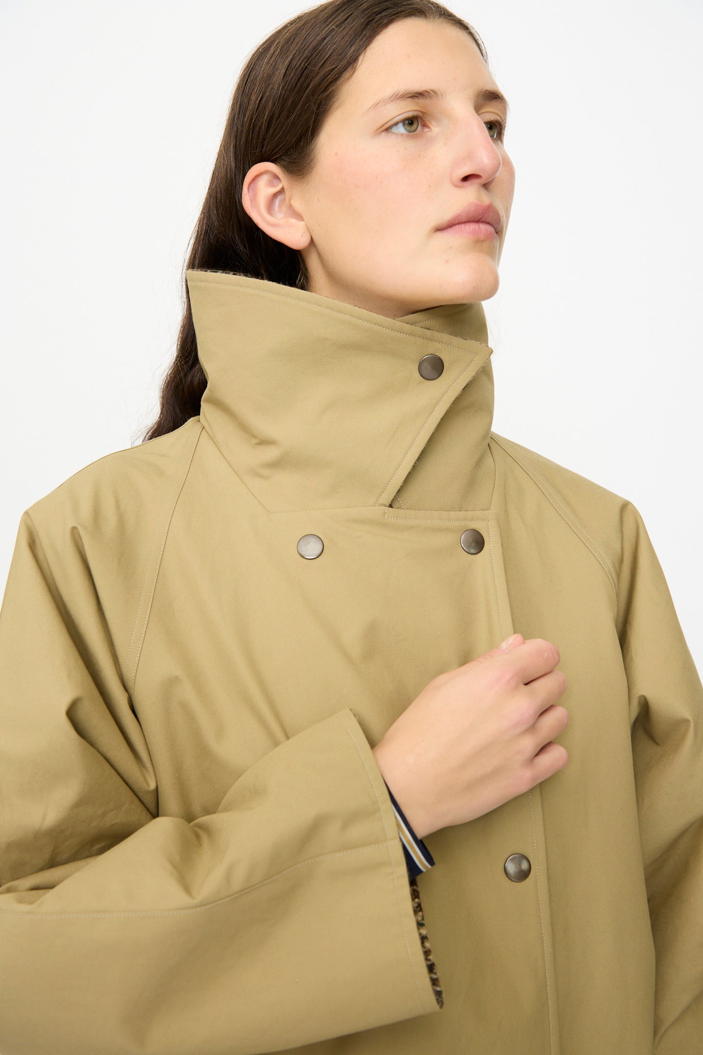 Model wearing the Les Prairies De Paris Dickens Moon Reversible British Trench in Taupe, featuring a high collar and snap buttons, looks off to the side against a plain white background.