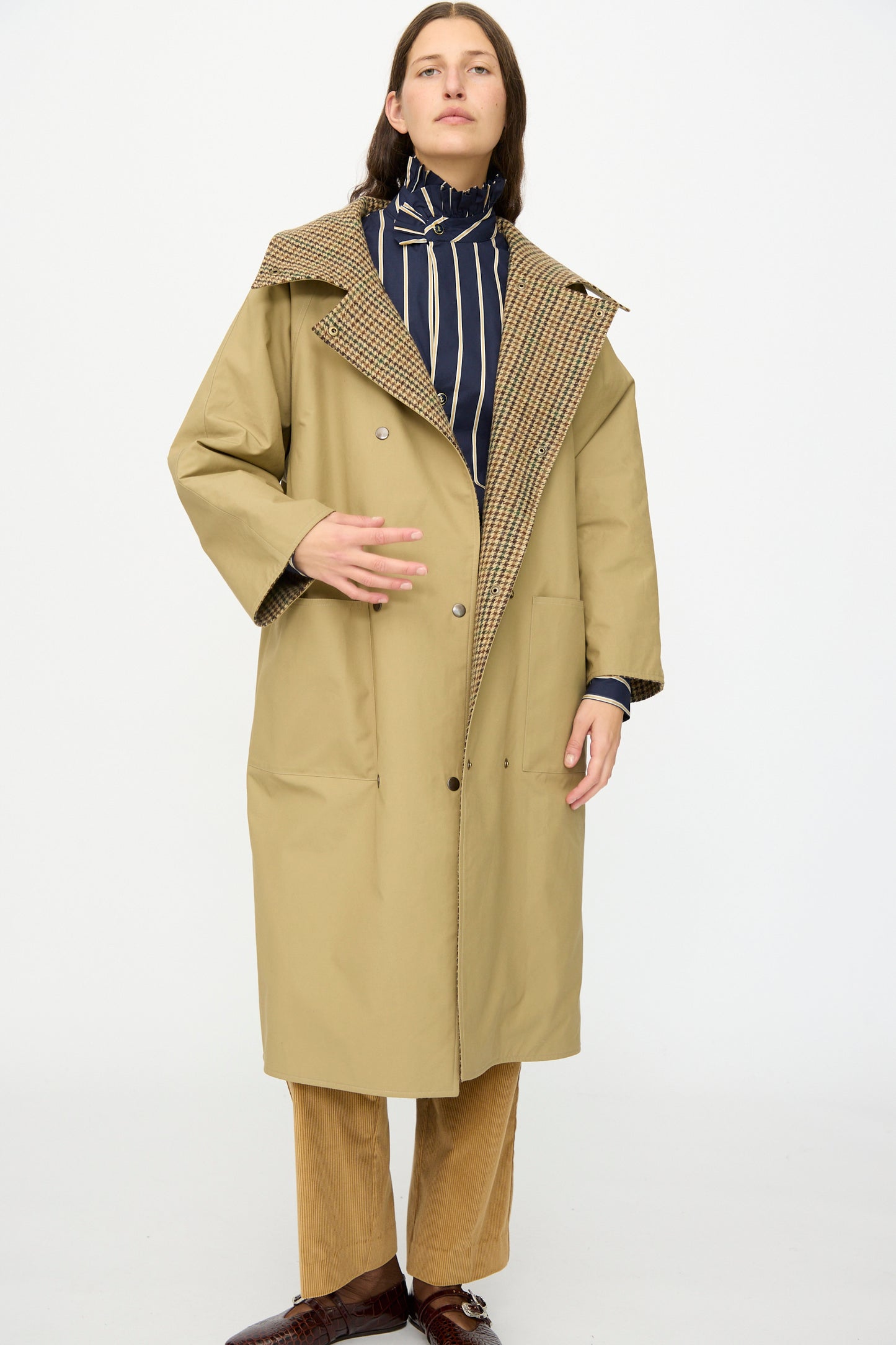 A person models the Les Prairies De Paris Dickens Moon Reversible British Trench in Taupe, featuring a patterned lining, layered over a striped high-neck blouse with brown pants and shoes against a plain white background.