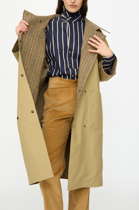 Someone wears the Les Prairies De Paris Dickens Moon Reversible British Trench in Taupe over a blue and white striped shirt and tan corduroy pants, holding it open to reveal its houndstooth wool lining.