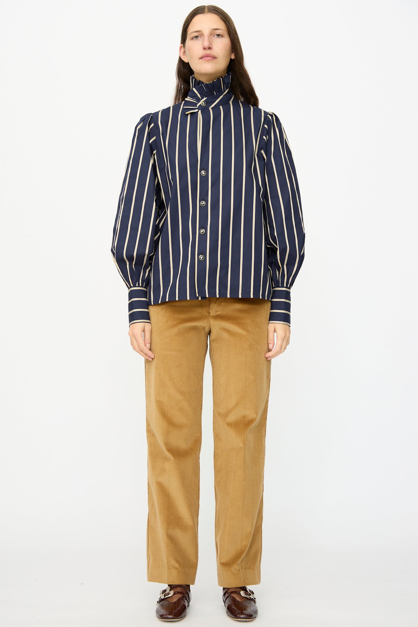 A person stands facing forward, wearing a navy and white vertical striped blouse, Gaoutcho Trouser in Miel by Les Prairies De Paris, and brown shoes, against a plain white background.