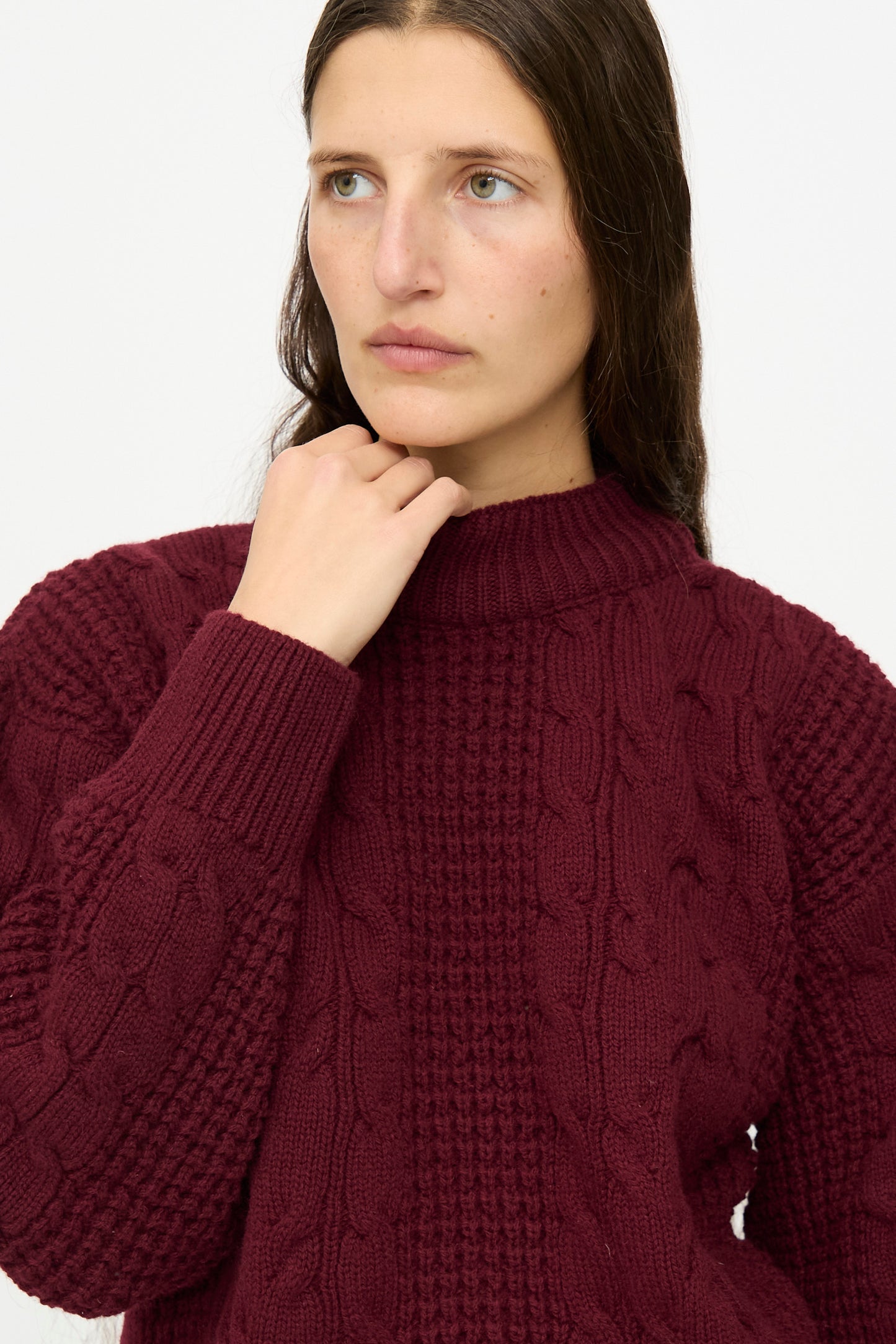 A person with long brown hair wears The Irish Wool Sweater in Bordeaux by Les Prairies De Paris, posing with one hand near their chin against a plain light background.