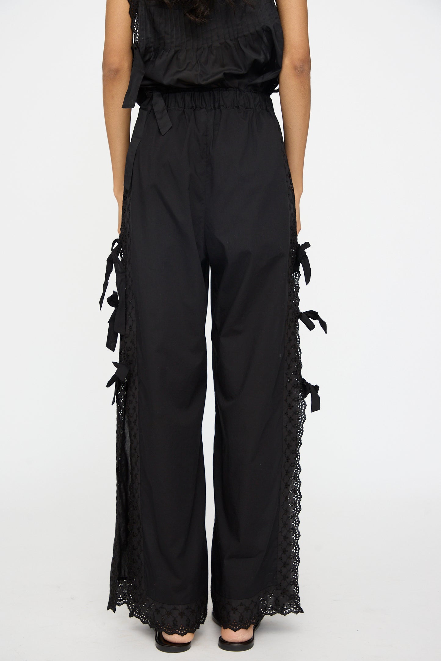 A person models the Clarissa Trousers in Nero by Loretta Caponi, featuring high-waisted black wide-leg pants with lace trim and bow details along the sides, made in Italy, viewed from the back against a plain white background.