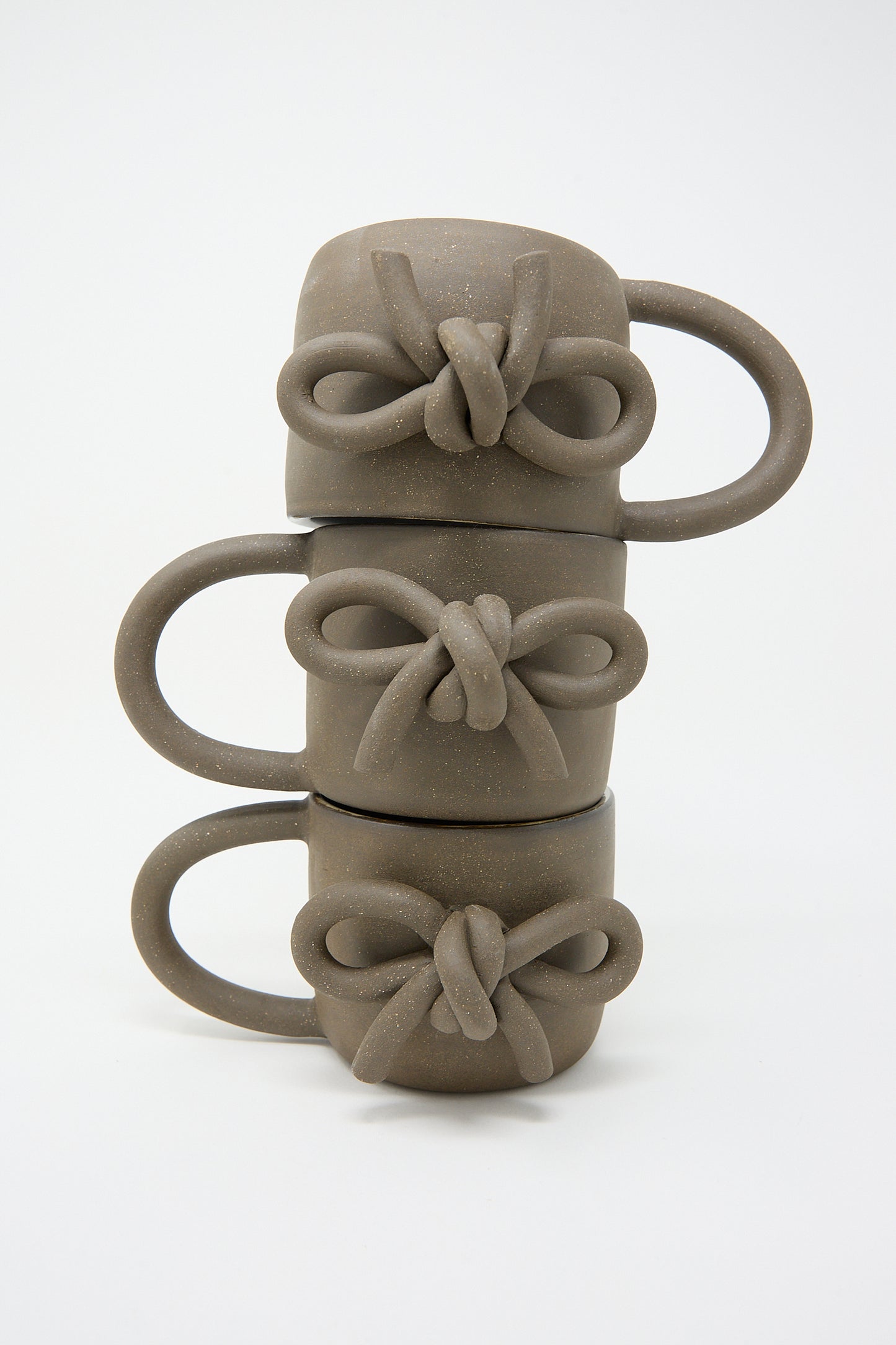 Four stacked Lost Quarry hand-formed ceramic mugs in black, each with a decorative bow design on the handle, against a white background.
