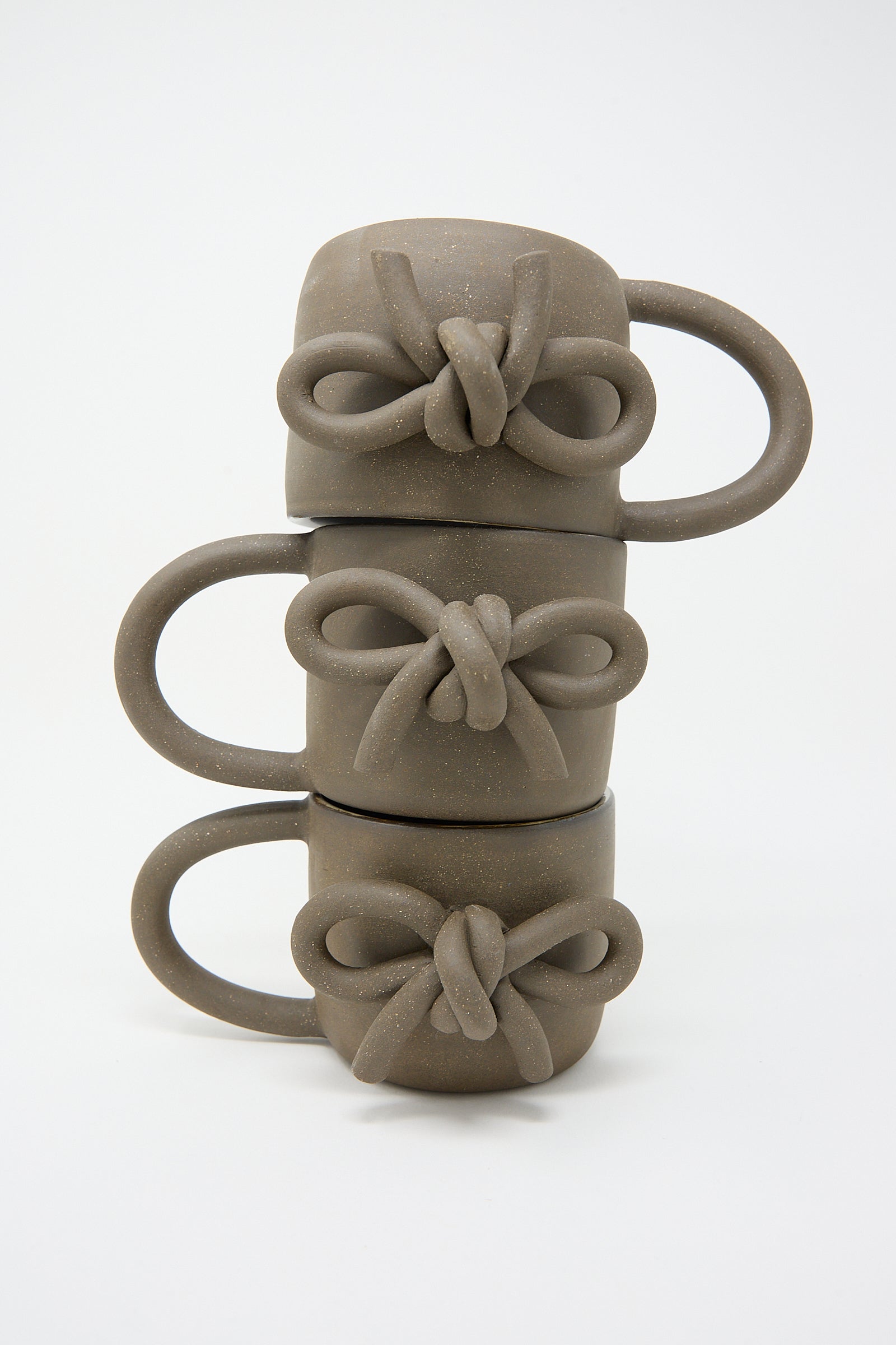 Four stacked Lost Quarry hand-formed ceramic mugs in black, each with a decorative bow design on the handle, against a white background.