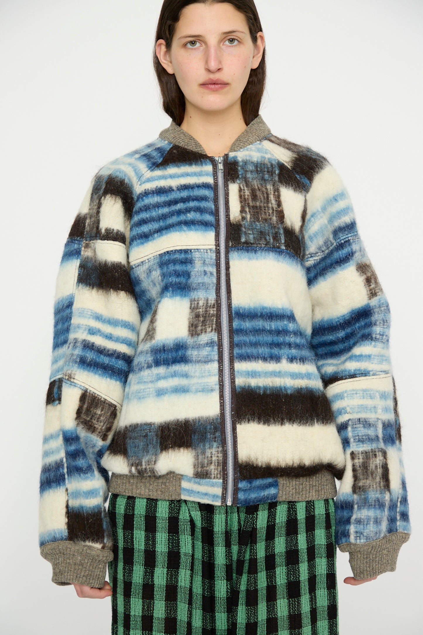 A person stands against a plain background, wearing the Luna Del Pinal Momostenango Woven Wool Blanket Bomber in Ocean over green plaid pants.