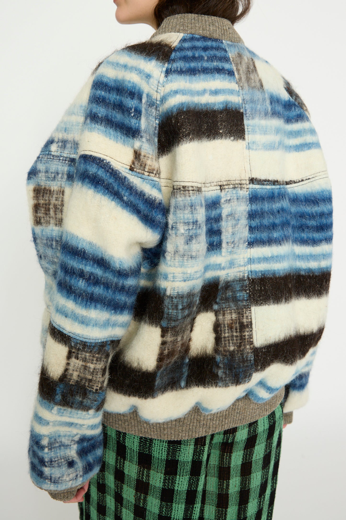 A person wears the Momostenango Woven Wool Blanket Bomber in Ocean by Luna Del Pinal, styled with a green and black checkered skirt.