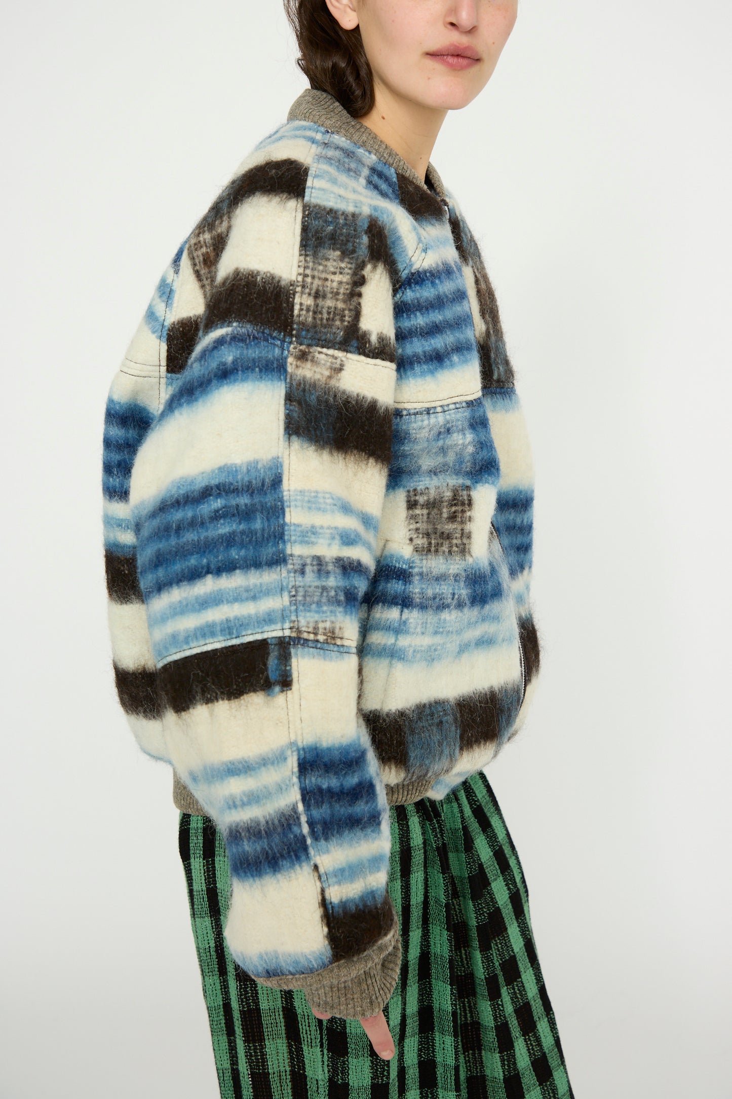 The person sports an oversized fit with Luna Del Pinal's Momostenango Woven Wool Blanket Bomber in Ocean, featuring brown accents, perfectly paired with green and black checkered wool pants.