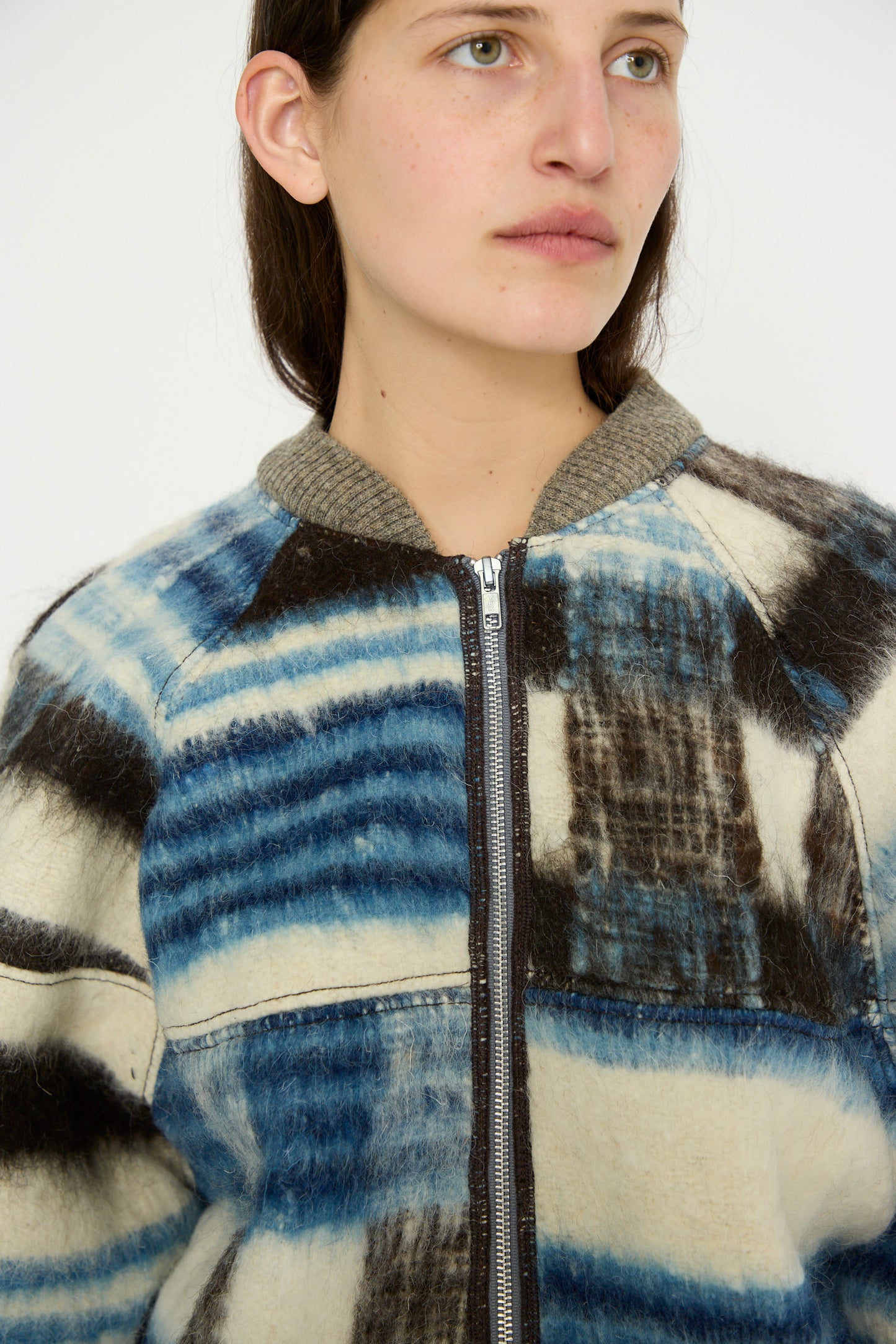 A person wearing the oversized Momostenango Woven Wool Blanket Bomber in Ocean by Luna Del Pinal, featuring a blue and brown plaid pattern with a silver zipper and gray collar.