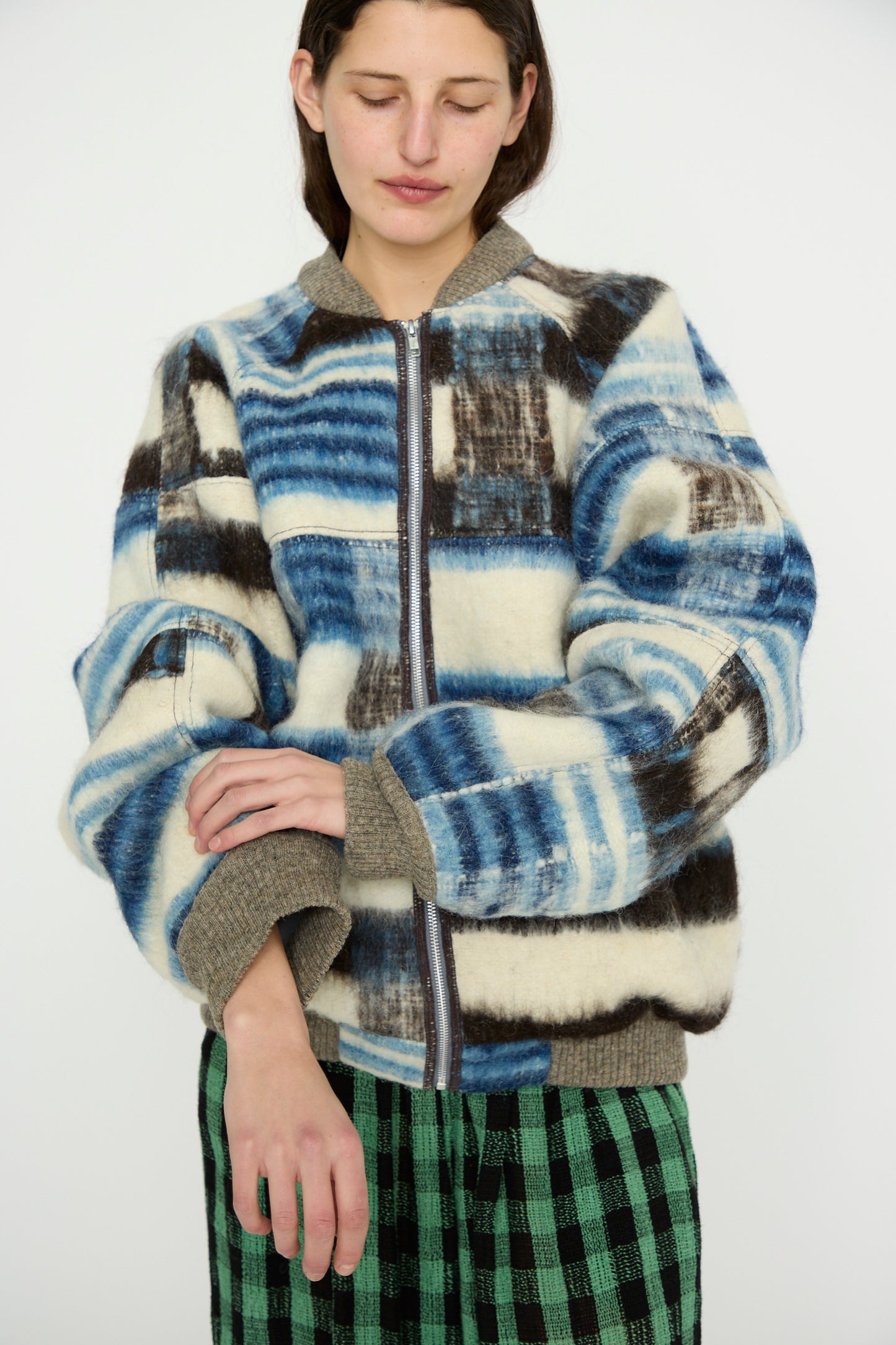 A person wearing the Momostenango Woven Wool Blanket Bomber in Ocean by Luna Del Pinal, featuring blue, black, and white patterns, pairs it with green checkered pants against a plain background.