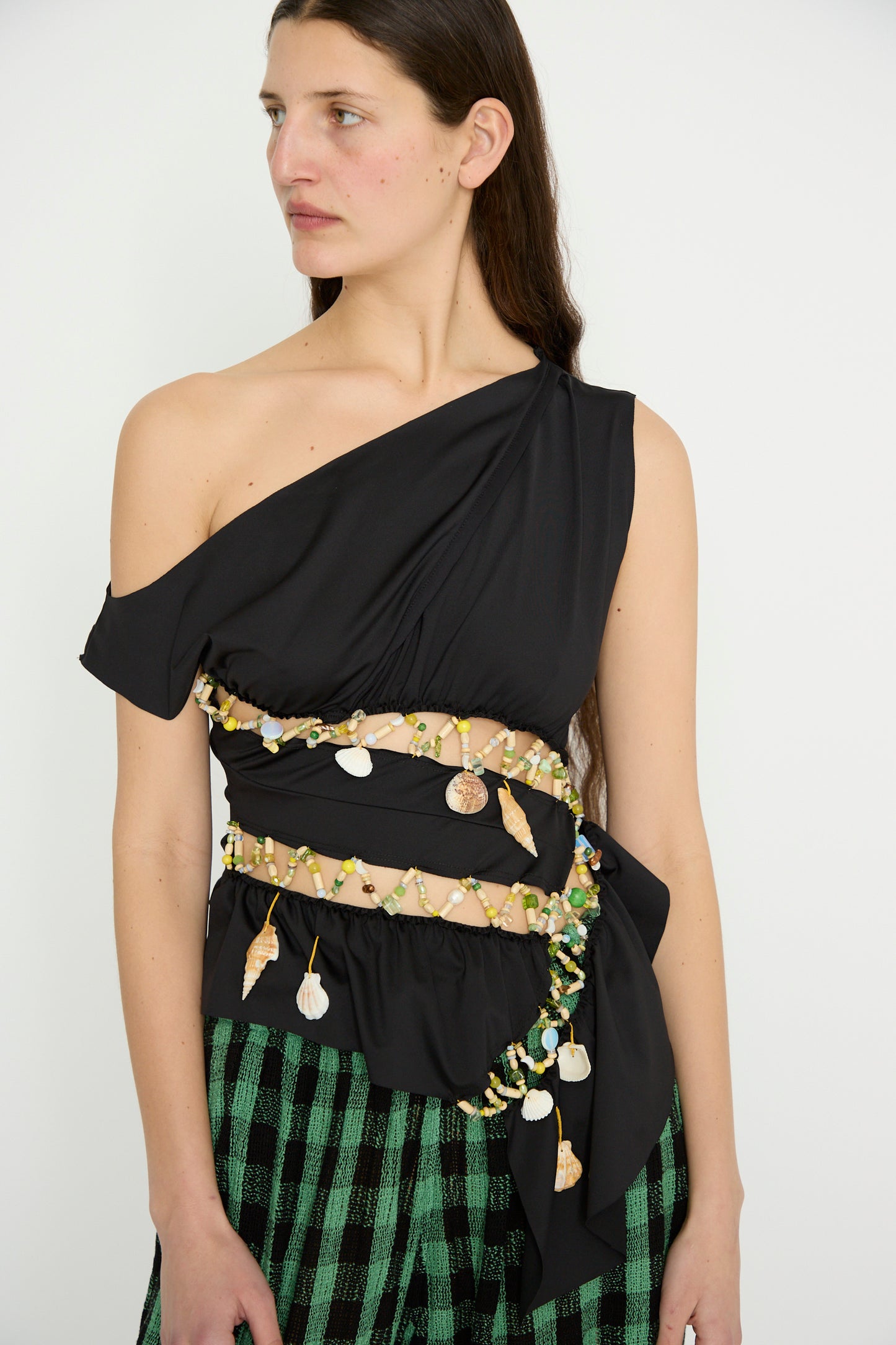 Person in a Luna Del Pinal Ornamental Off Shoulder T-Shirt in Night Sea, featuring shell and bead decorations, paired with green checkered pants made of recycled polyester, looking to the side.
