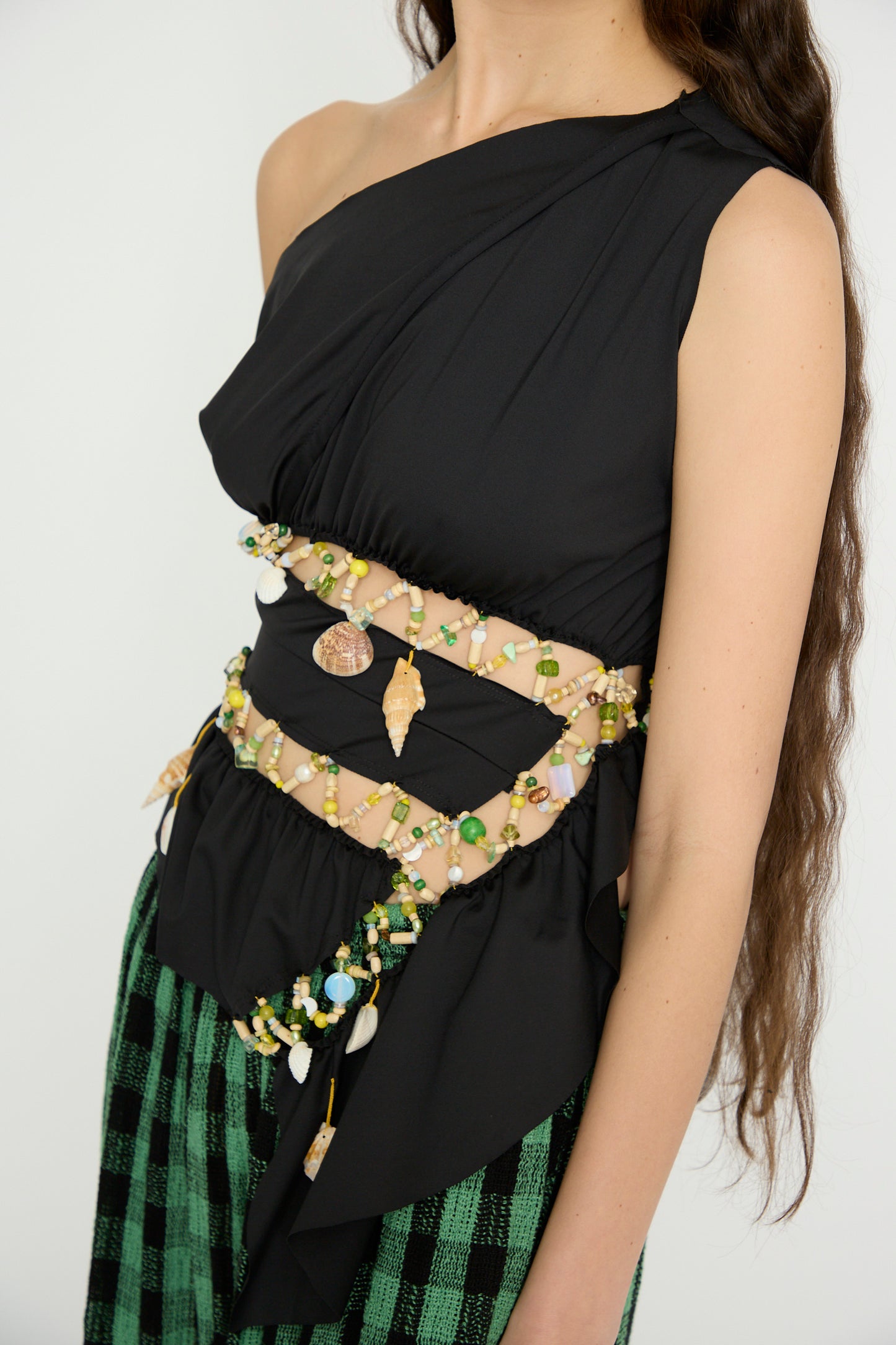 A person wearing Luna Del Pinal's Ornamental Off Shoulder T-Shirt in Night Sea and green checkered pants, adorned with elaborate beaded waist and arm chains.
