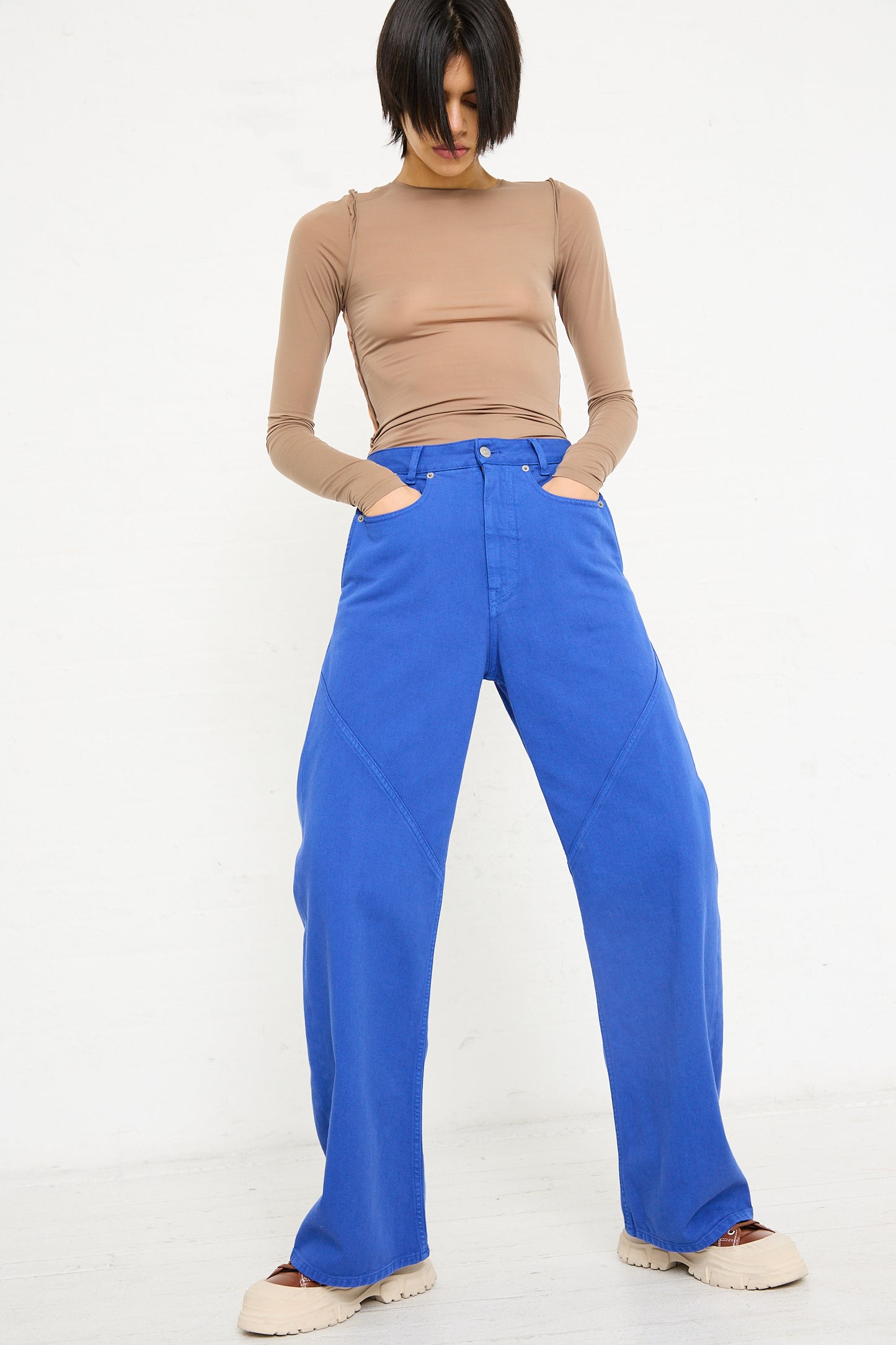 Woman standing in a studio, wearing a tan top and blue MM6 5 Pocket Pant high-waisted trousers, with her hands in pockets.