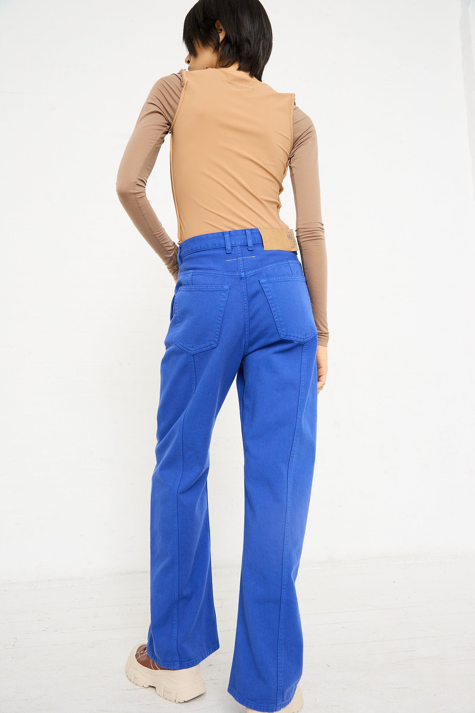 A woman from behind, wearing the 5 Pocket Pant in Blue by MM6 and a beige long-sleeve top, standing against a white wall.