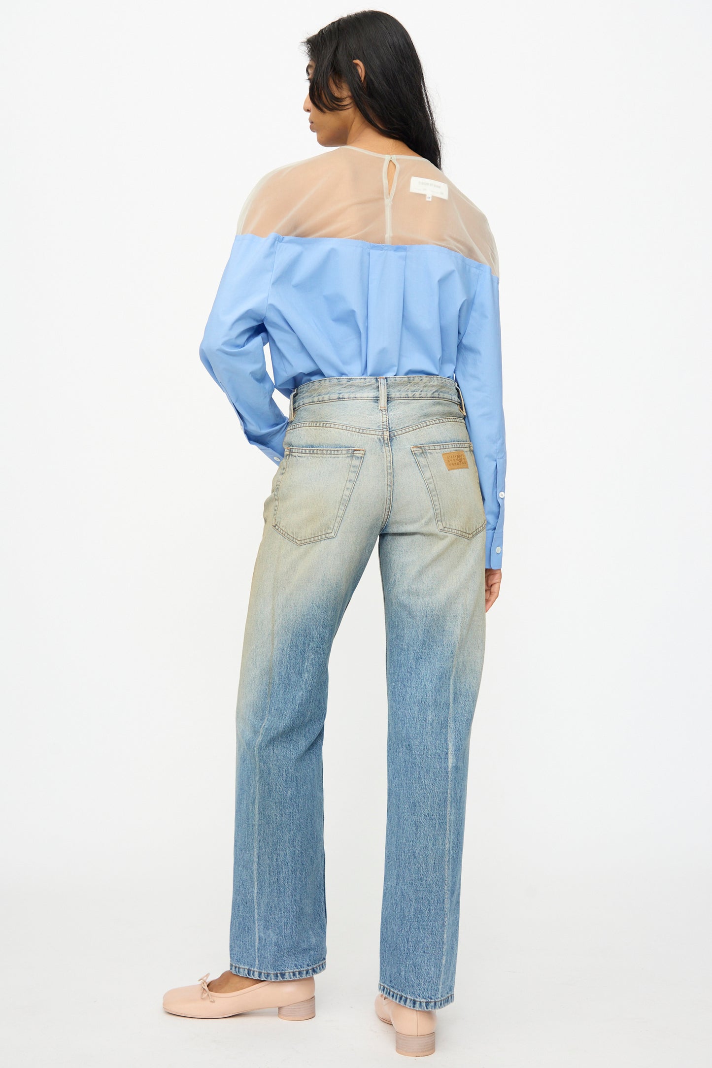 A person stands facing away, wearing a blue shirt with a sheer beige yoke, MM6 5 Pockets Pants in Medium Blue, and light pink shoes.
