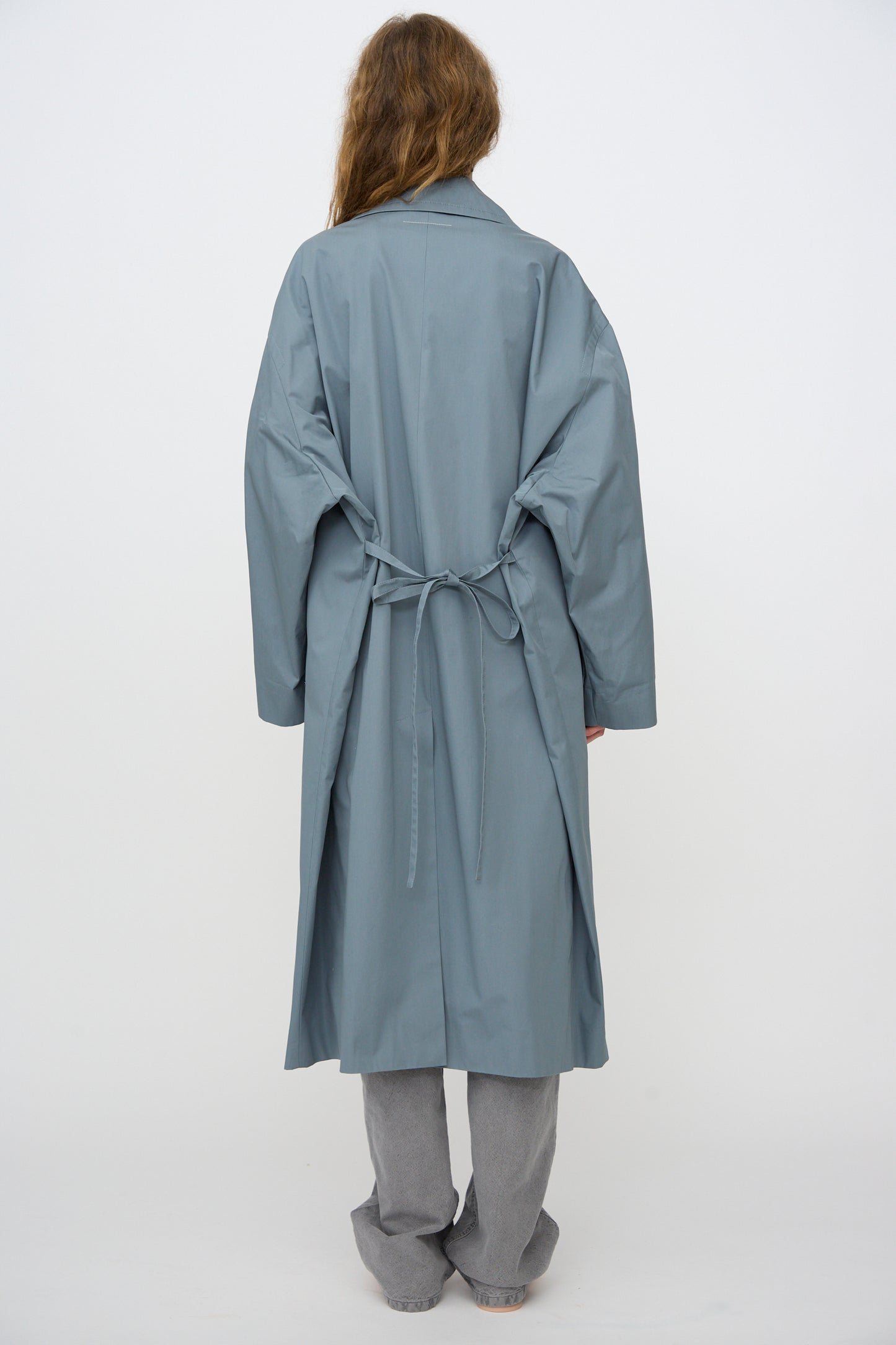 Person stands with back to camera, wearing the MM6 Coat in Grey Blue—an oversized, water-repellent cotton style tied at the waist—paired with gray pants.
