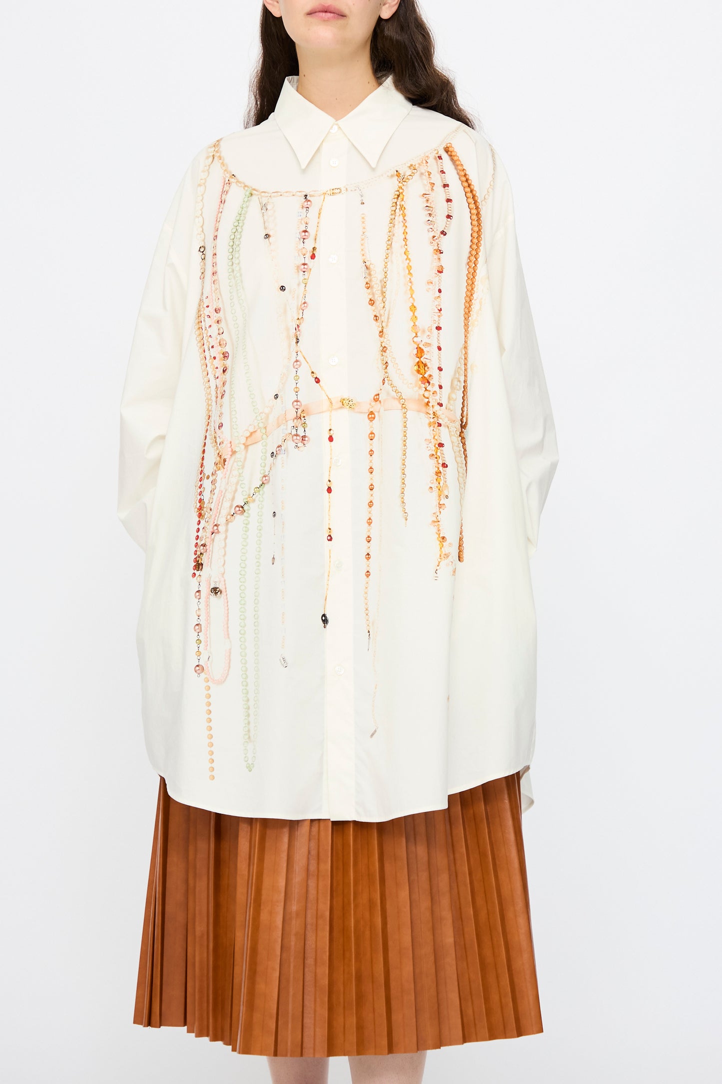 A woman wears the MM6 Dress in Pearls Print, an oversized white button-up shirt featuring bead and chain details, paired with a brown pleated skirt.