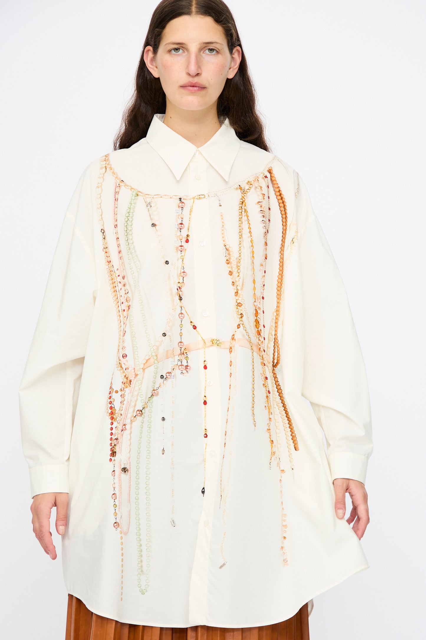 A woman wears the MM6 Dress in Pearls Print, an oversized white button-up shirt dress featuring a vivid pearl-inspired motif, standing against a plain background.