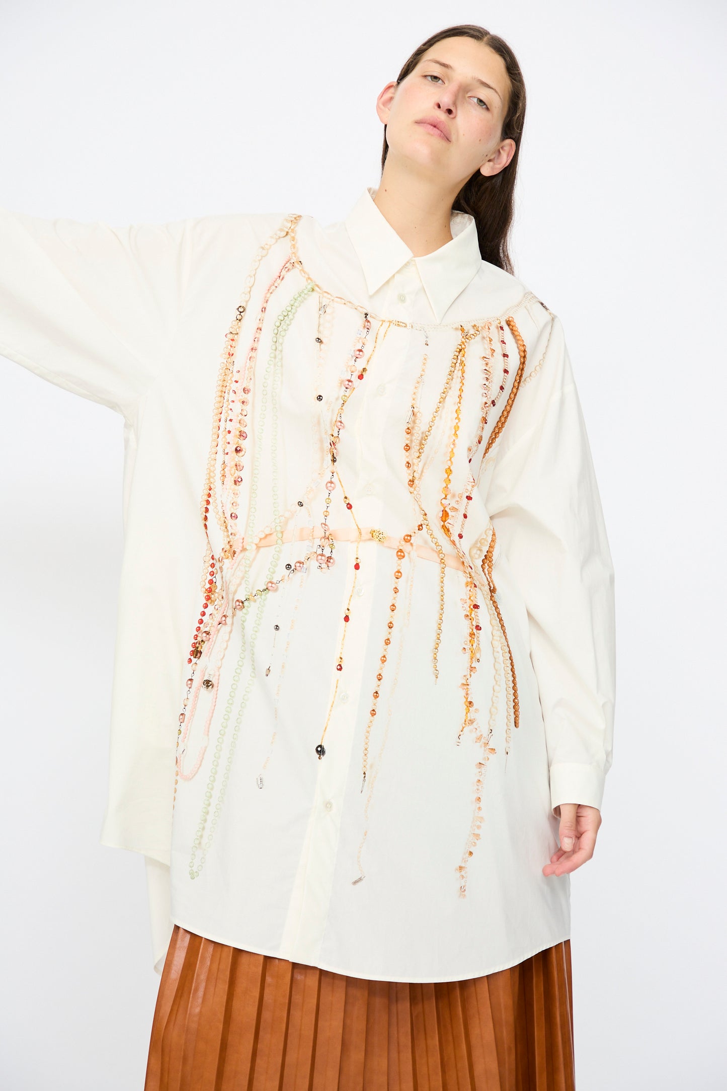 A person wears the MM6 Dress in Pearls Print, featuring an oversized fit and pearl chain detailing, styled with a pleated brown skirt and posed against a plain white background.