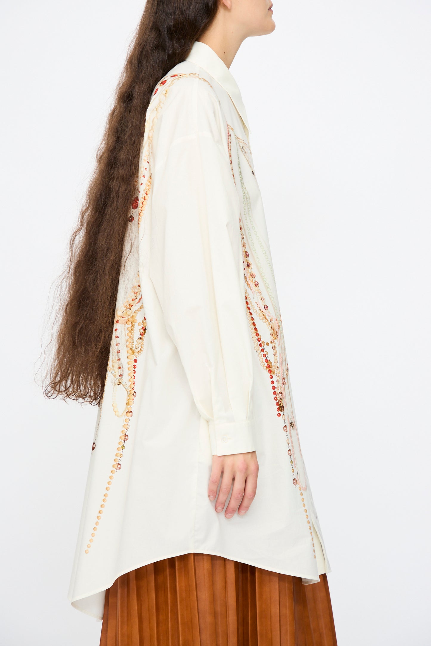 A person with long brown hair is seen from the side wearing the MM6 Dress in Pearls Print—a cream-colored, oversized cotton shirt dress with a decorative chain motif—paired with a pleated brown skirt.