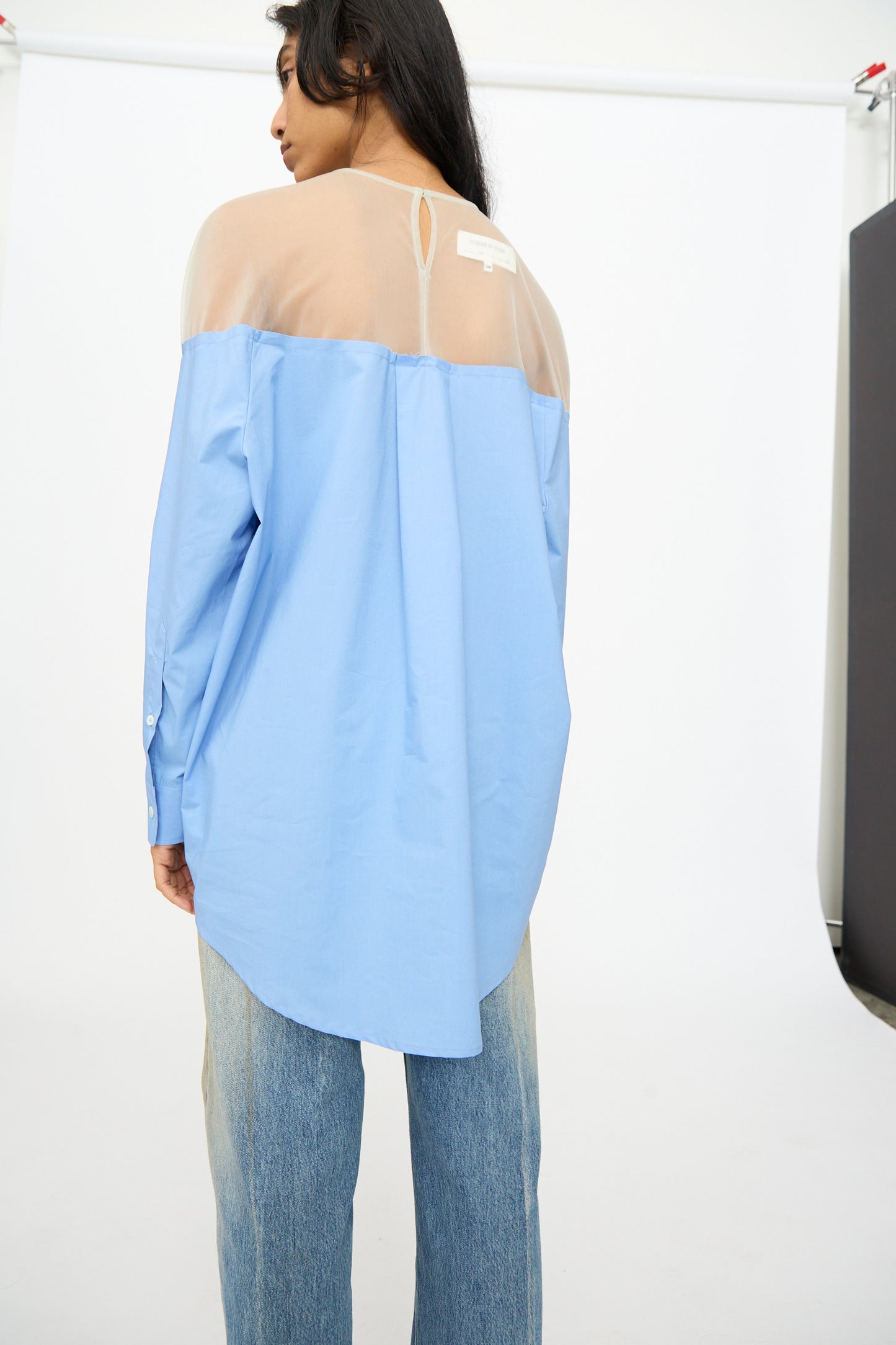 A person with their back to the camera wears the MM6 Long Sleeved Top in Sky Blue, featuring a sheer upper section, paired with light blue jeans. Photo taken against a plain white background.