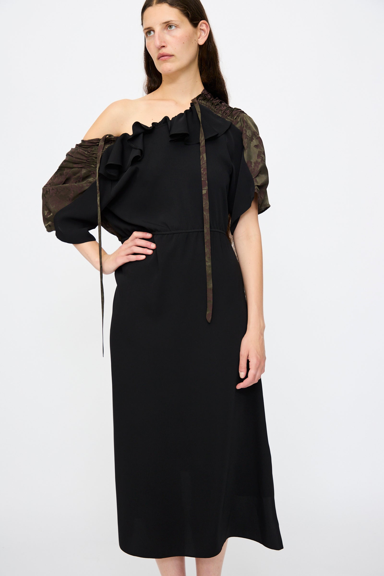 A woman stands against a plain white background wearing the MM6 Midi Dress in Black and Khaki Burgundy, featuring a relaxed fit, asymmetrical off-shoulder ruffled neckline, and one camouflage-patterned sleeve.