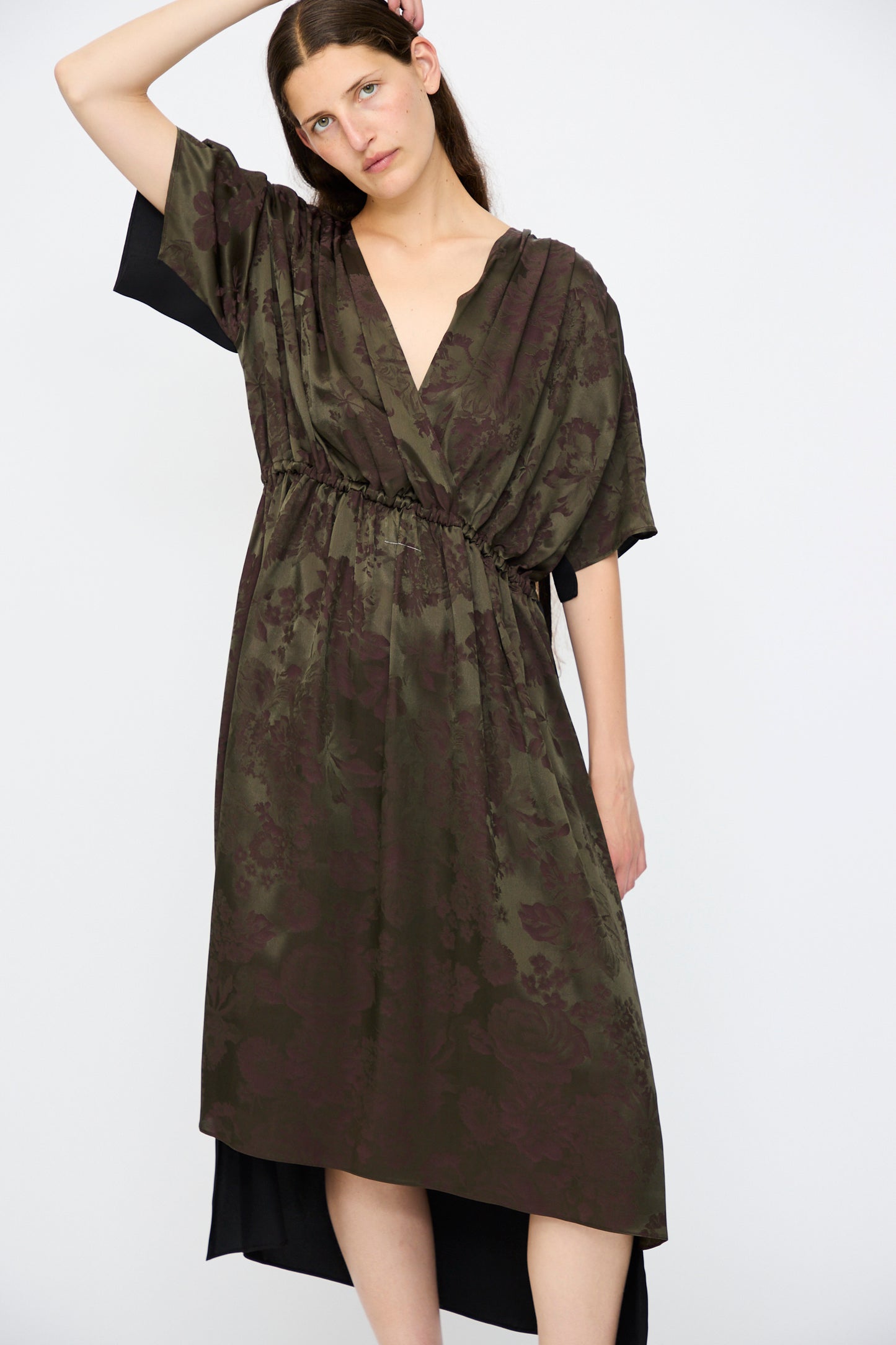 A woman with long brown hair wears the MM6 Midi Dress in Black and Khaki Burgundy, a relaxed-fit, floral-print wrap dress with short sleeves, standing against a plain white background.