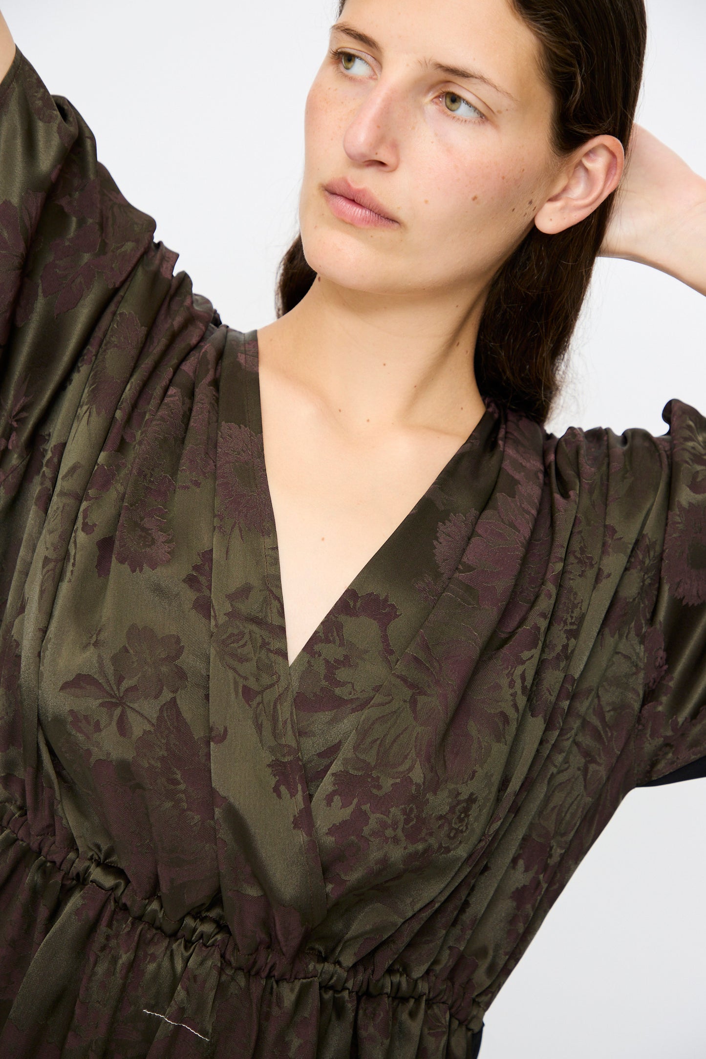 A person wearing the MM6 Midi Dress in Black and Khaki Burgundy, featuring a relaxed fit and deep V-neckline, looks to the side with one arm raised.